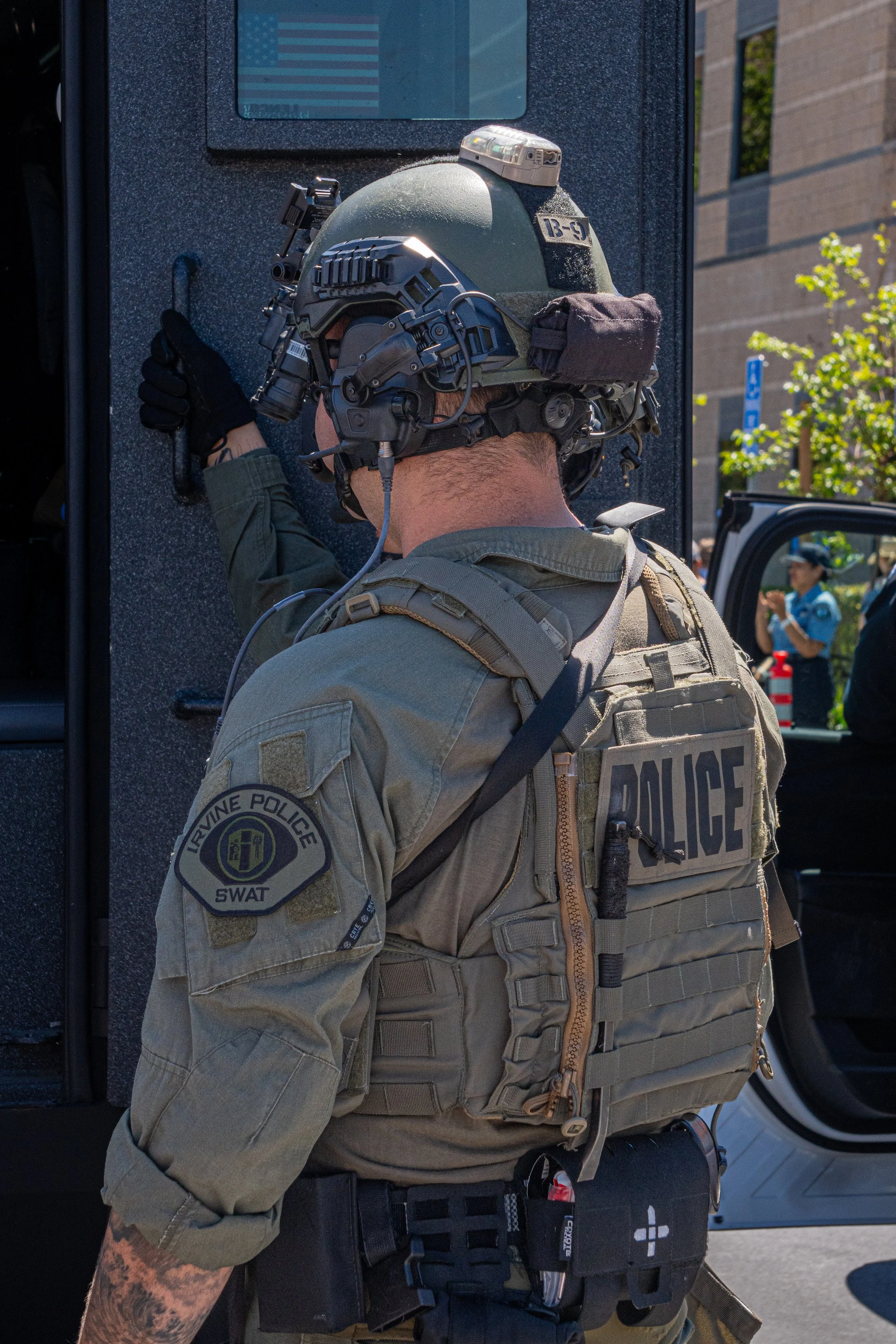 A police officer wearing tactical gear and a helmet with communication equipment, leaning against a vehicle during what appears to be a police operation or event.