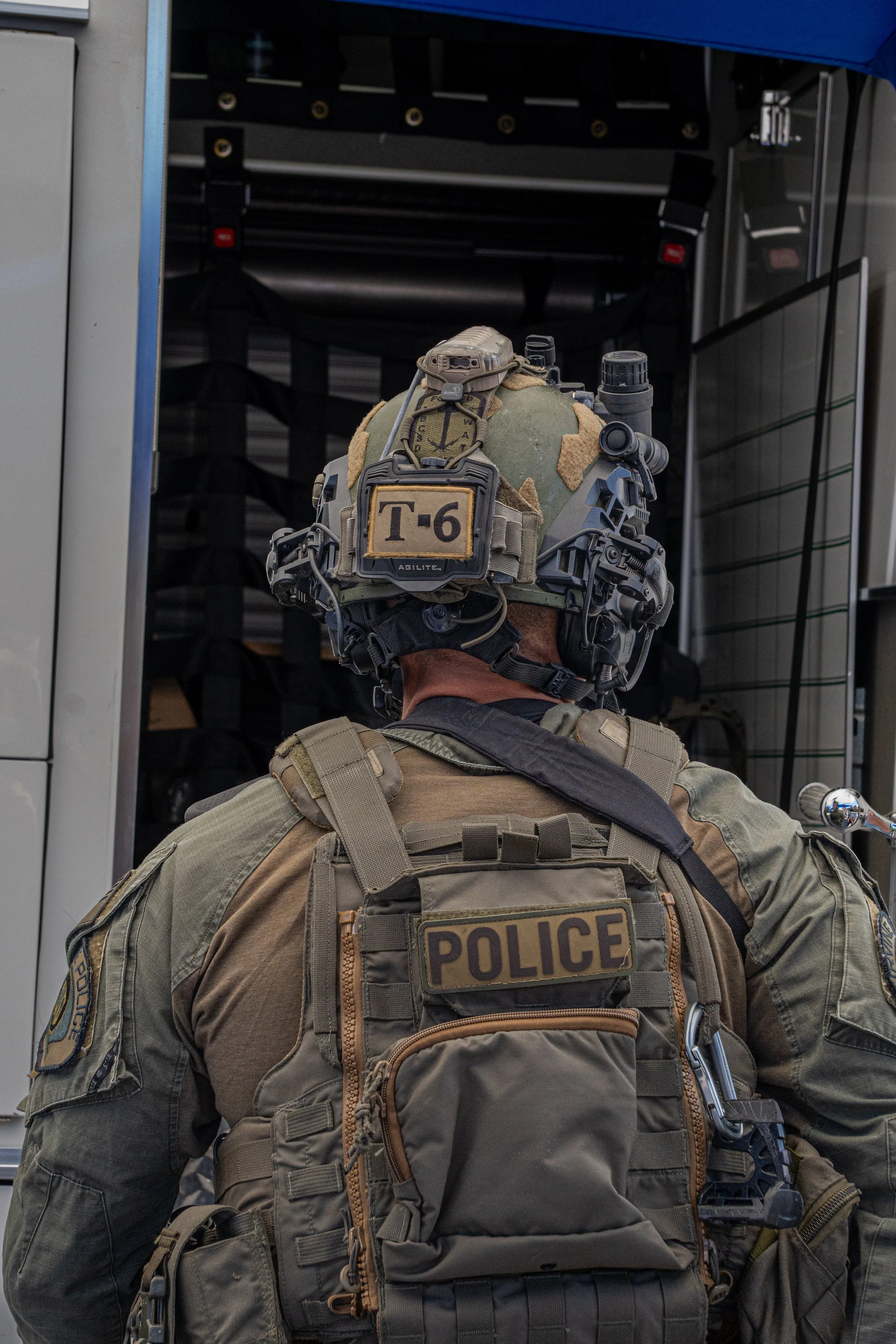 A police officer in tactical gear, including a helmet with electronics, facing away, standing in front of a police vehicle.