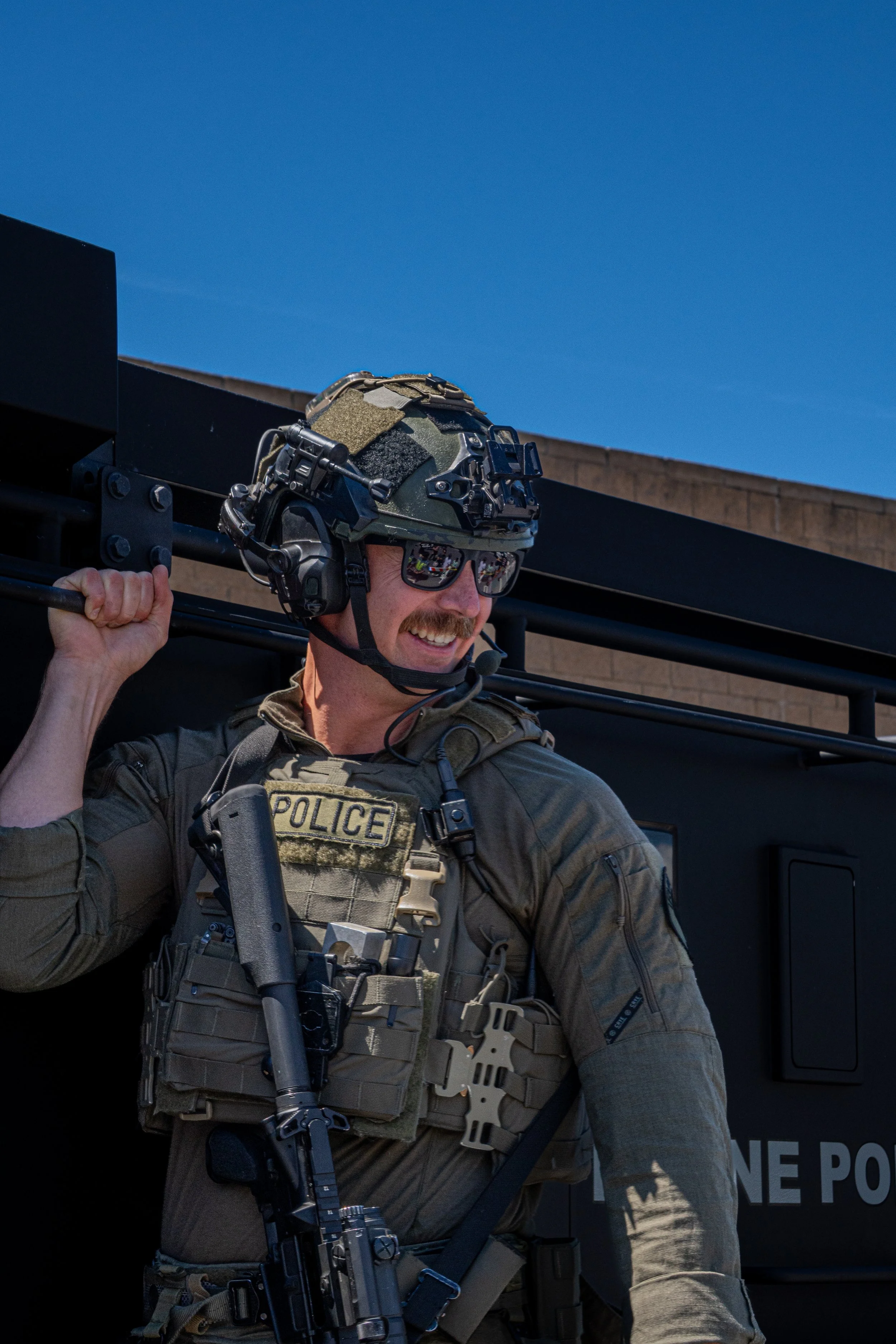 A police officer wearing tactical gear, including a helmet and sunglasses, smiling and leaning against a military-style vehicle under a clear blue sky.