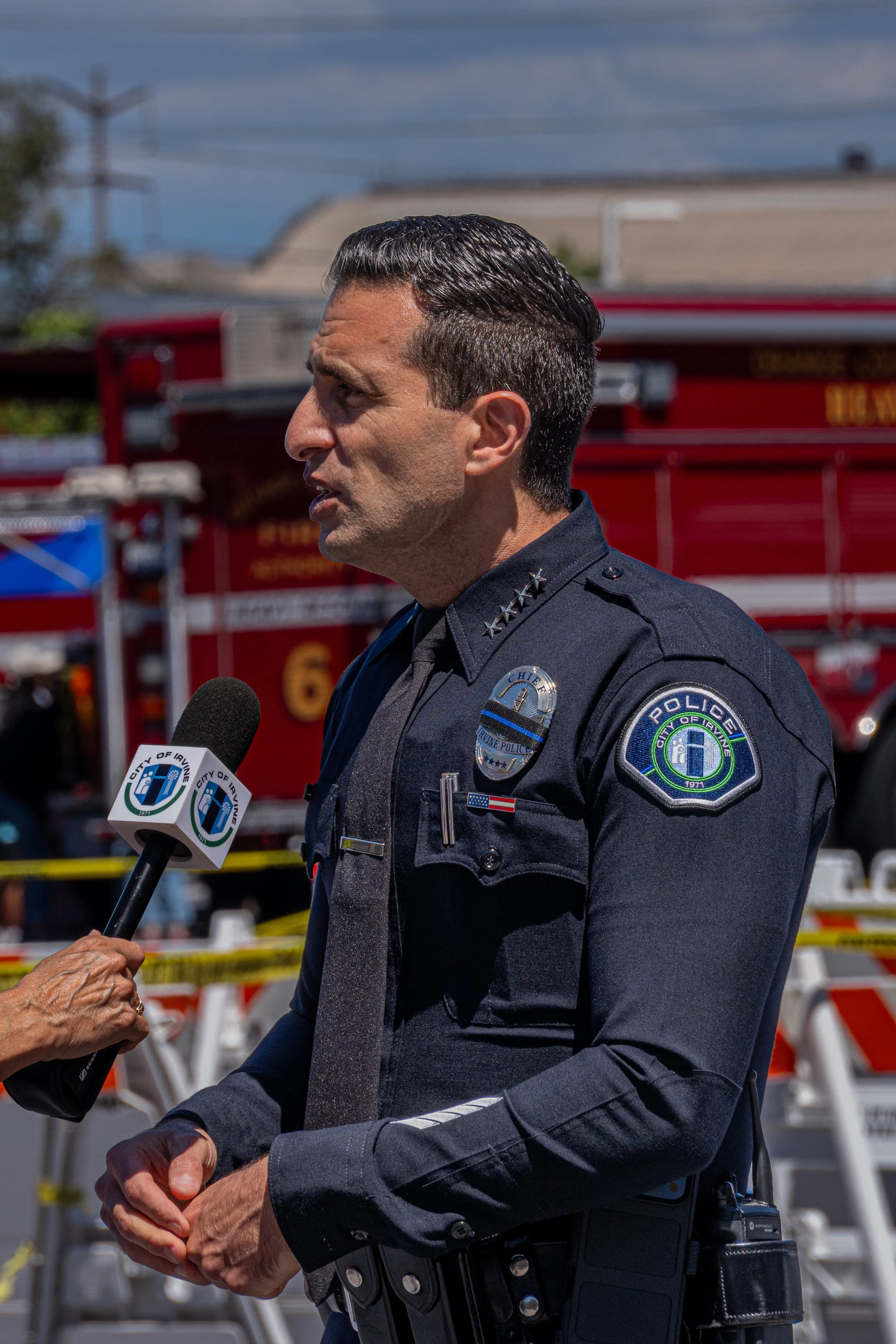 A police officer speaking into a microphone during a press conference, with fire trucks and caution tape in the background.