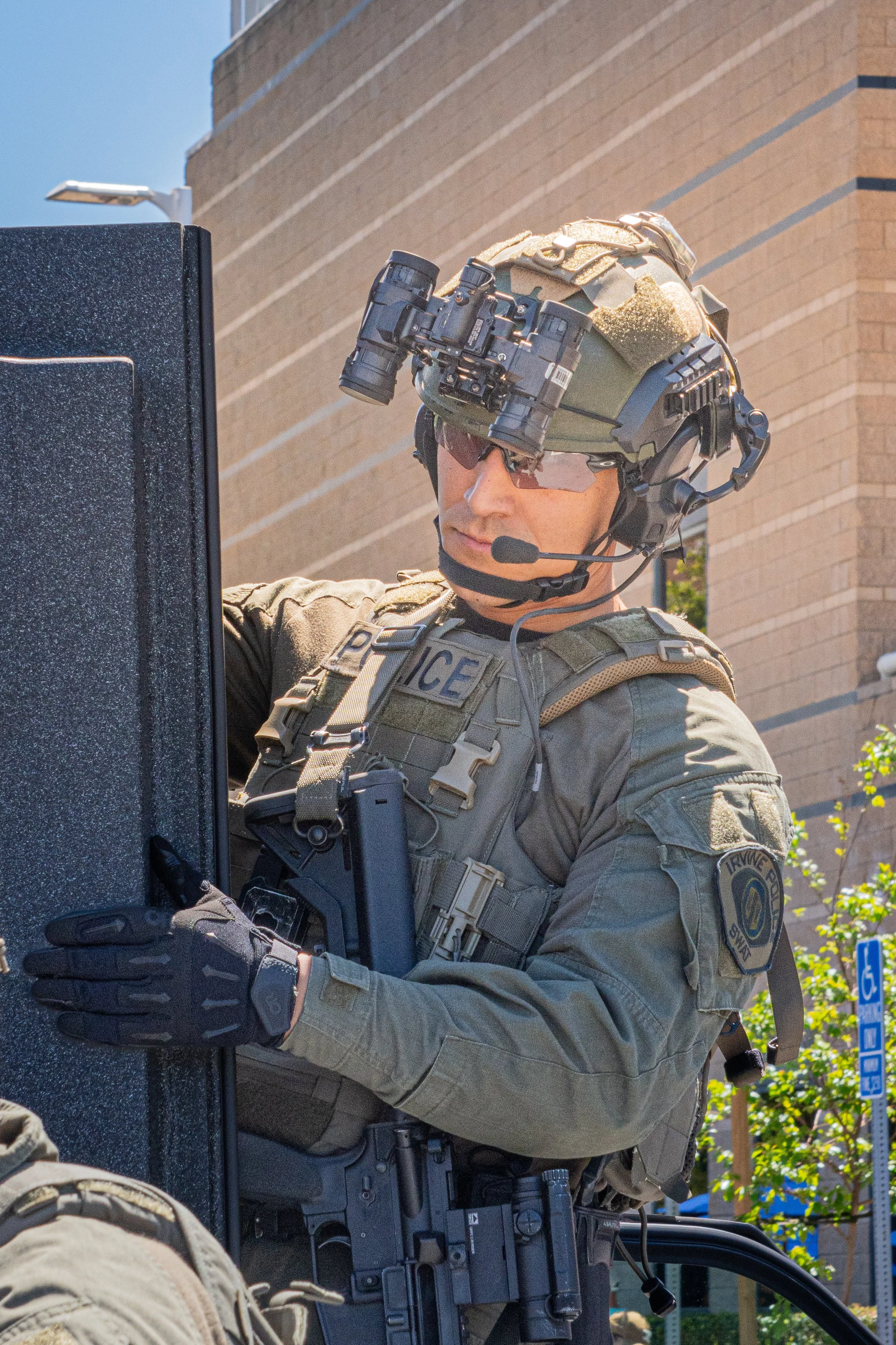 A police officer equipped with tactical gear, including a helmet with mounted night vision goggles, sunglasses, a headset with microphone, and body armor, working on a computer outdoors in front of a brick building.