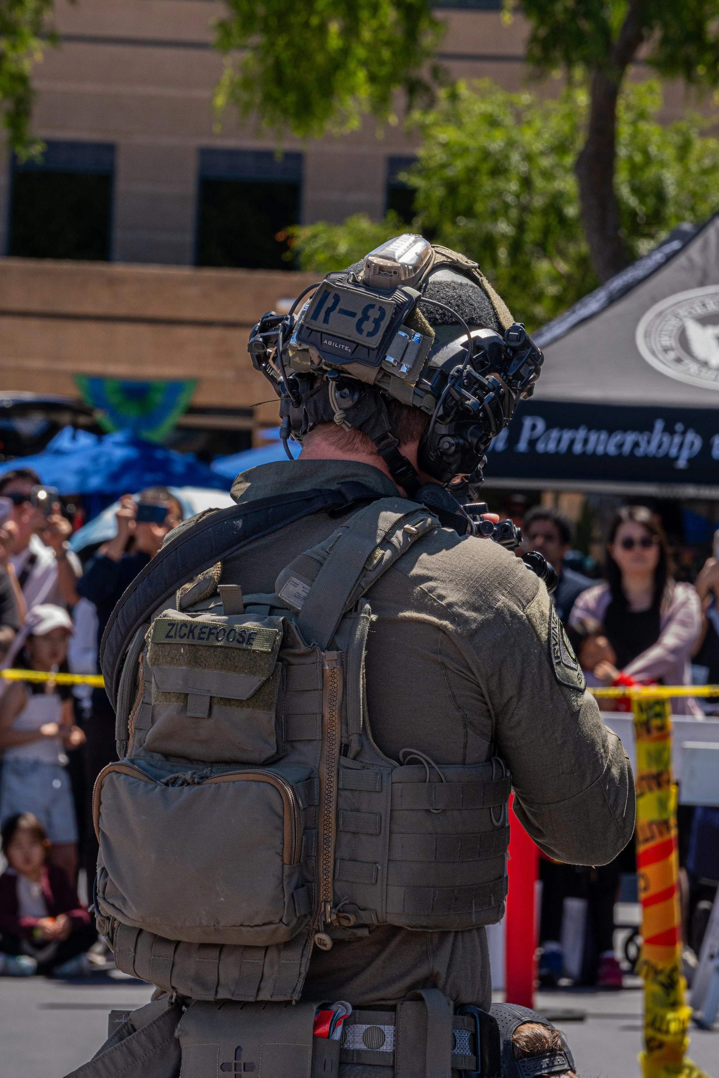 A military or tactical personnel wearing a helmet and tactical gear, standing outdoors in front of a crowd of people at an event or protest.