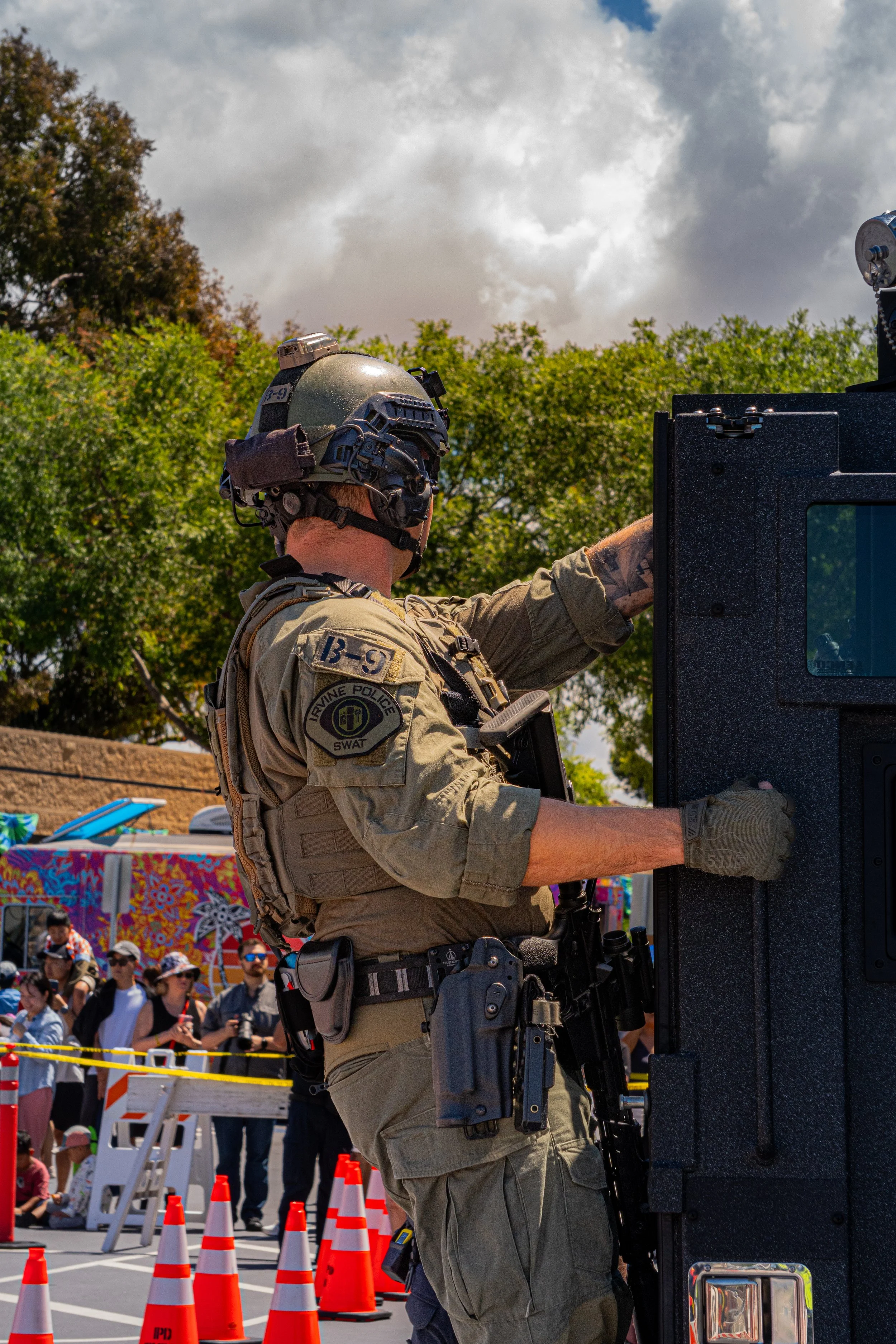 A police officer wearing a helmet and tactical gear interacts with a large black equipment or vehicle at an outdoor public event, with spectators and colorful tents in the background.