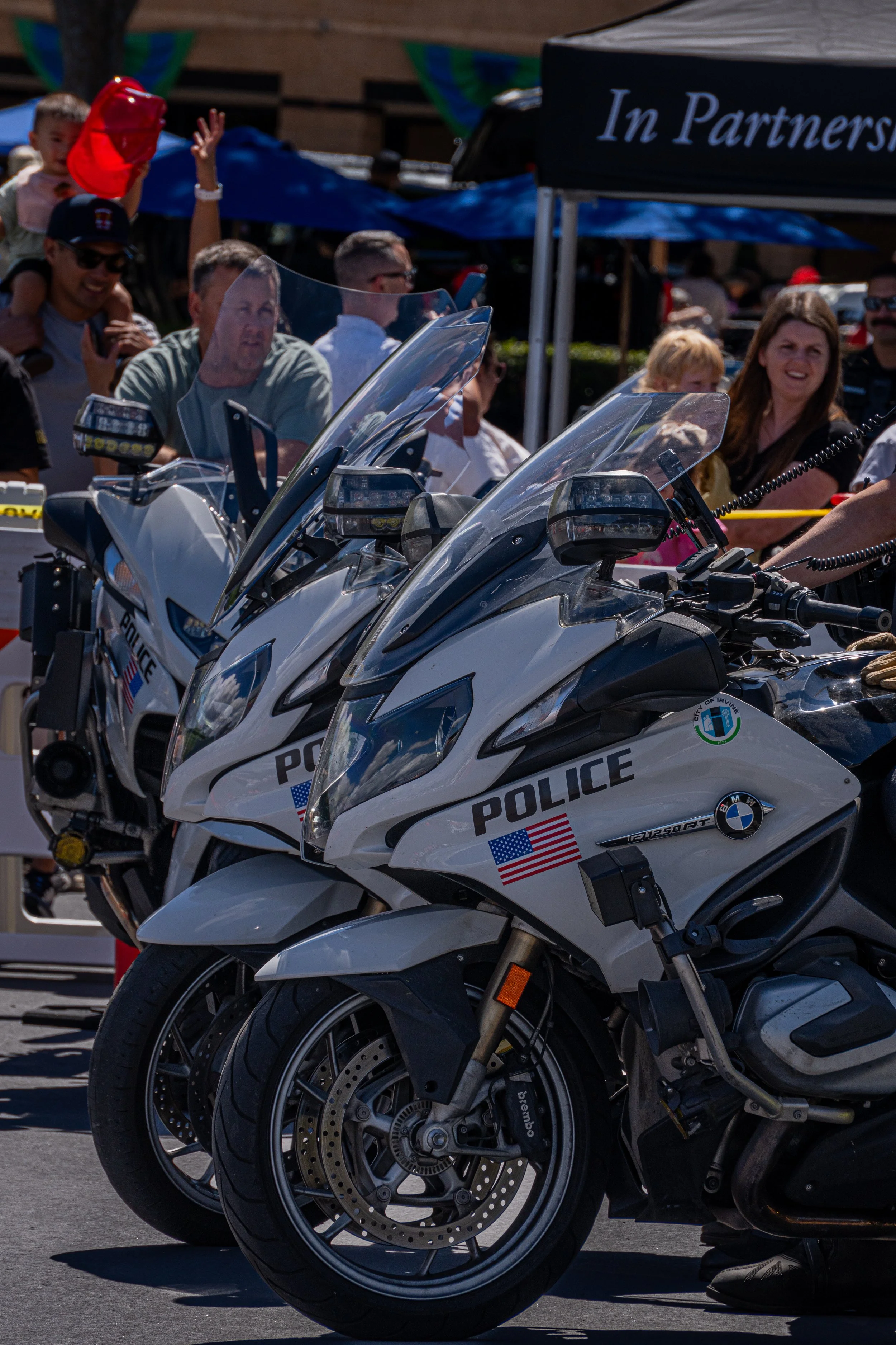 Two police motorcycles parked in a row at a public event, with a crowd of people and tents in the background.
