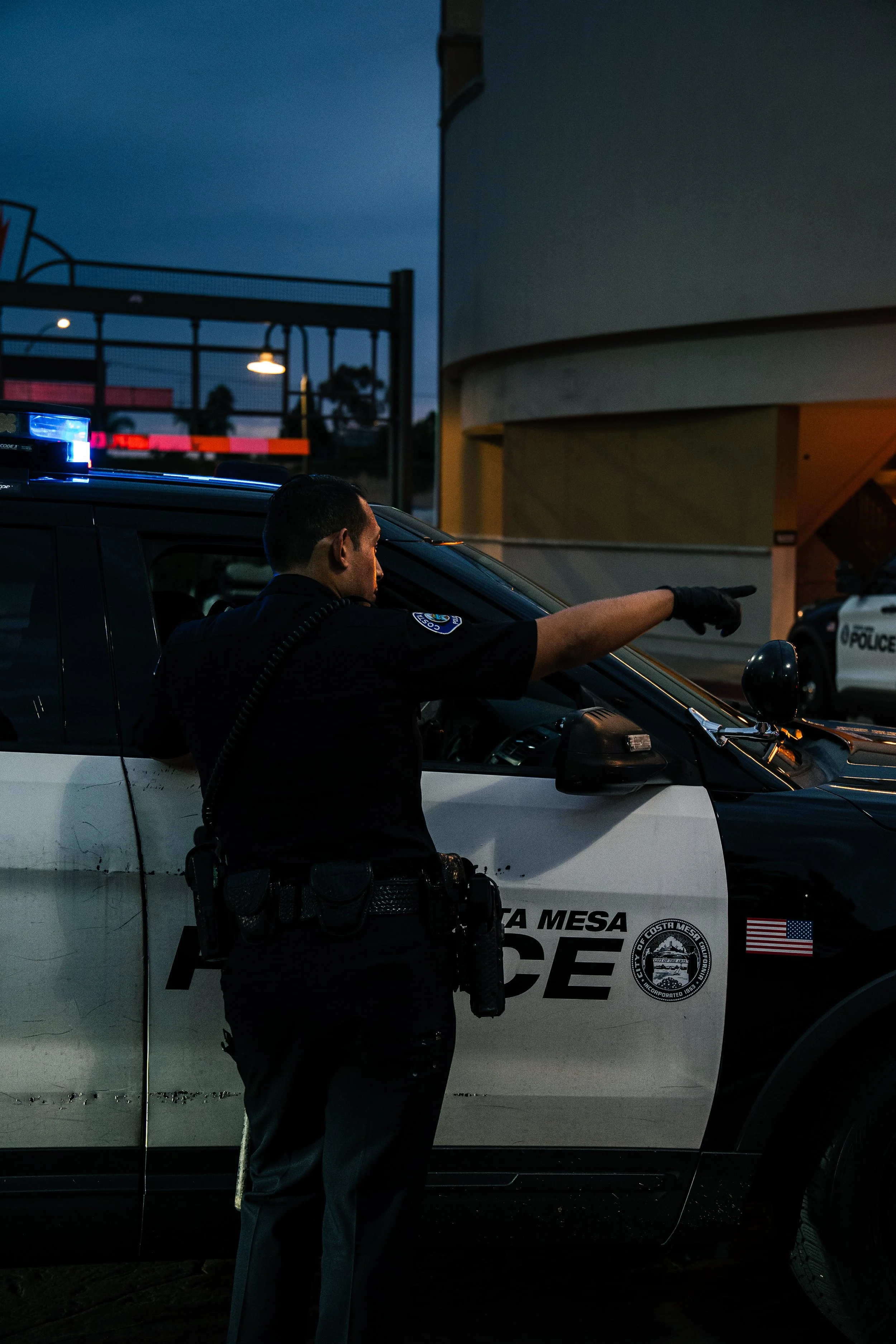 A police officer in a dark uniform is gesturing with his right arm while standing next to a police vehicle, which is parked outside near a building during evening or night. The police vehicle has a light bar on the roof and markings that include the 
