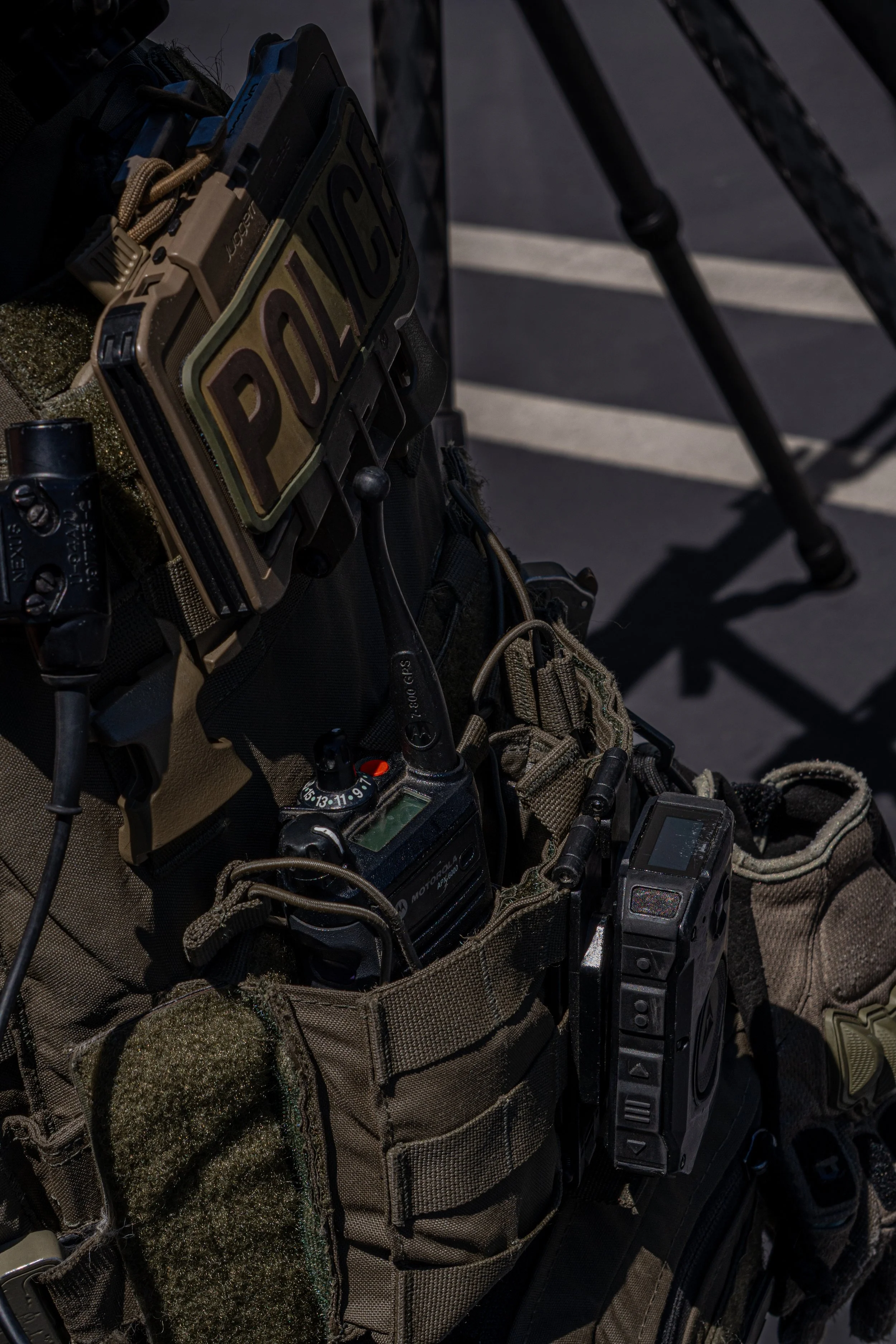 Close-up of tactical police gear including a vest with various electronic devices, a radio, and a patch labeled 'POLICE' attached to the vest.