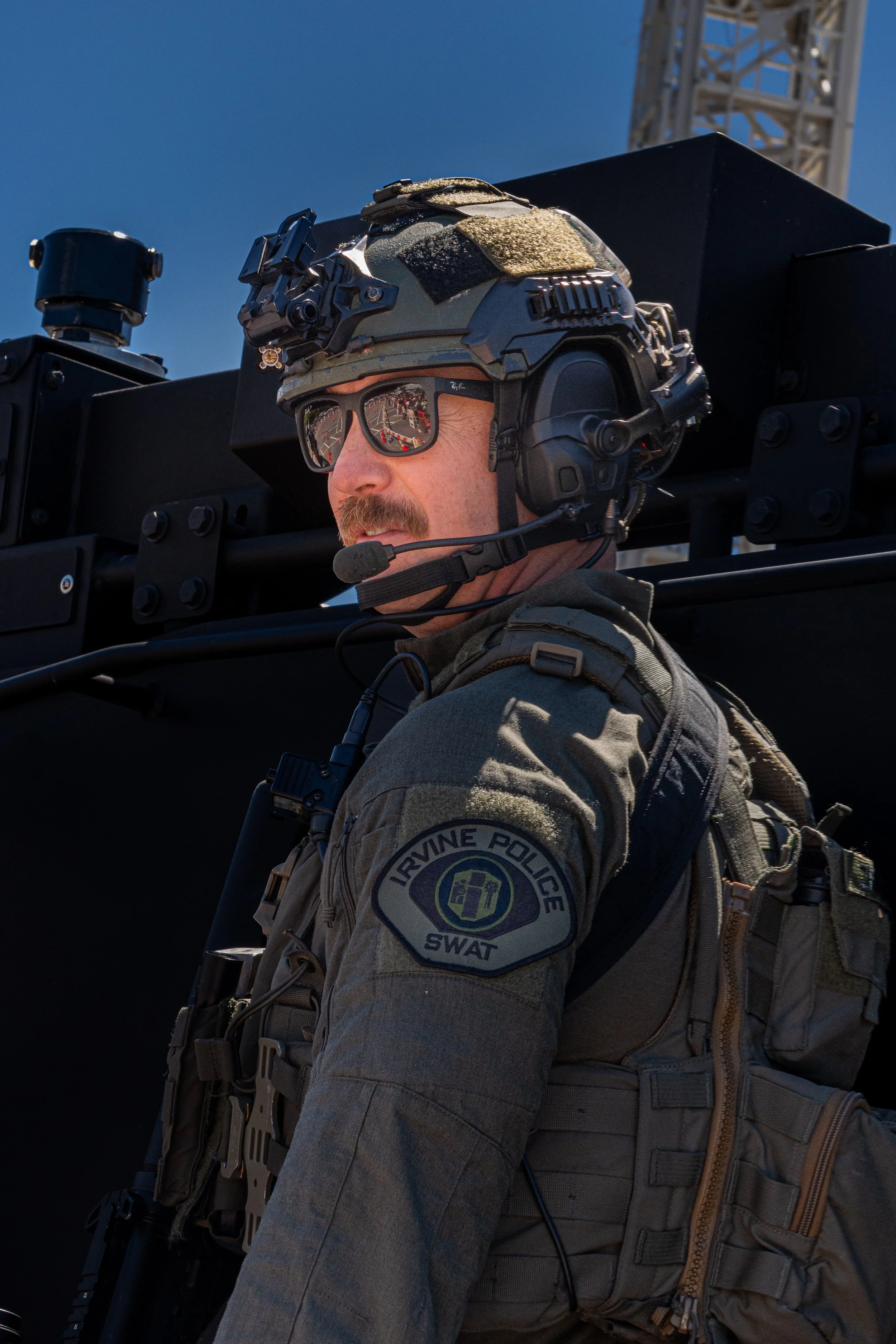 A soldier in tactical gear and sunglasses, wearing a helmet with mounted equipment, is standing outdoors against a clear blue sky, with a large black military vehicle behind him.