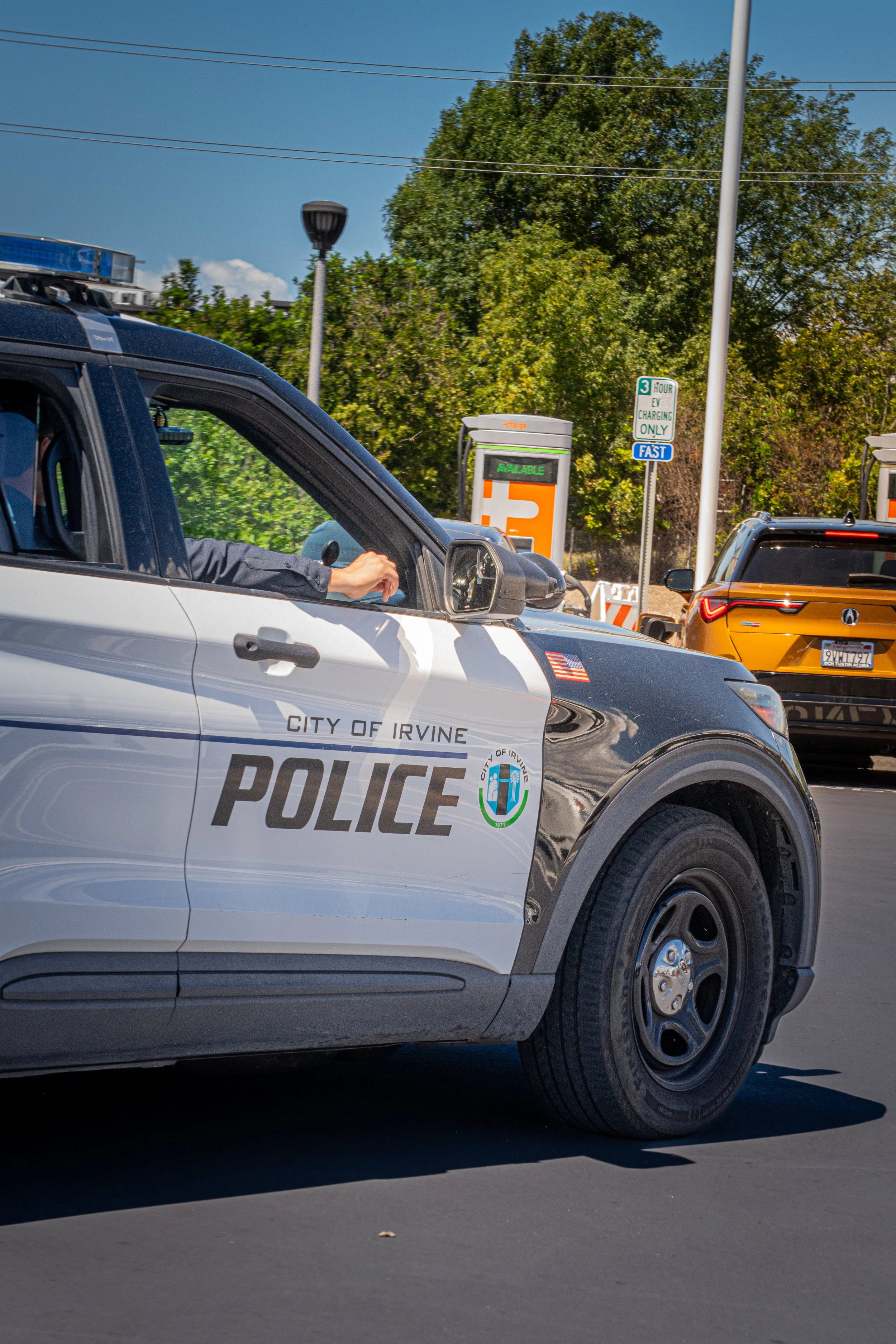 A City of Irvine police car on a city street with a driver inside. There are trees, a lamp post, and parking meters in the background.