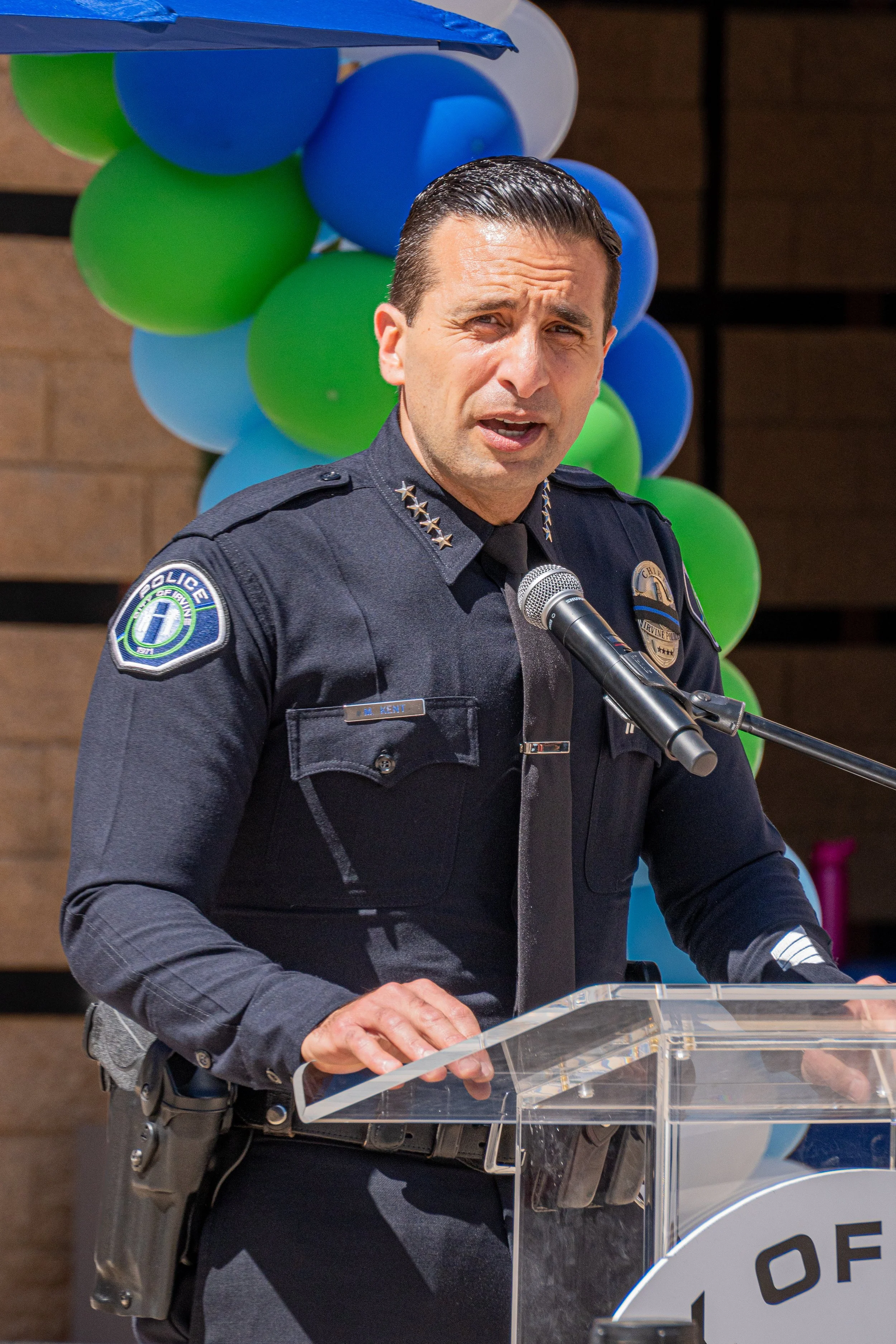 A police officer speaking at a podium with a backdrop of blue, green, and light blue balloons.