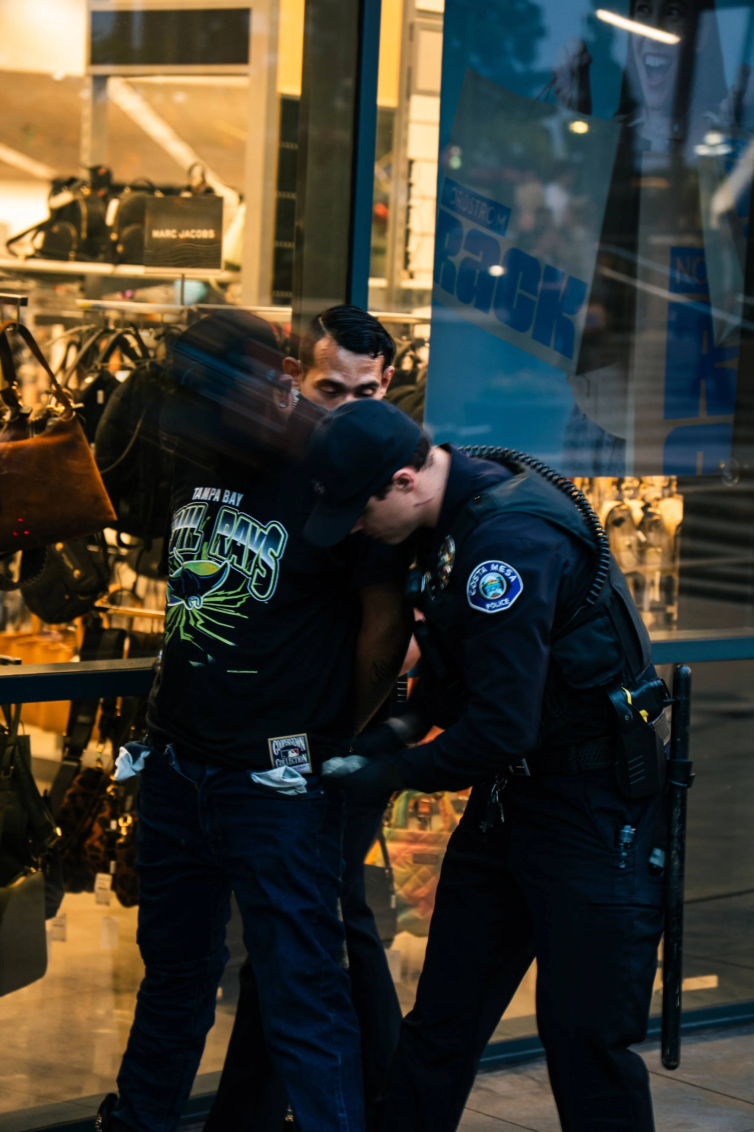 Two police officers are detaining a man outside a retail store, with security cameras and shelves of bags visible in the background.