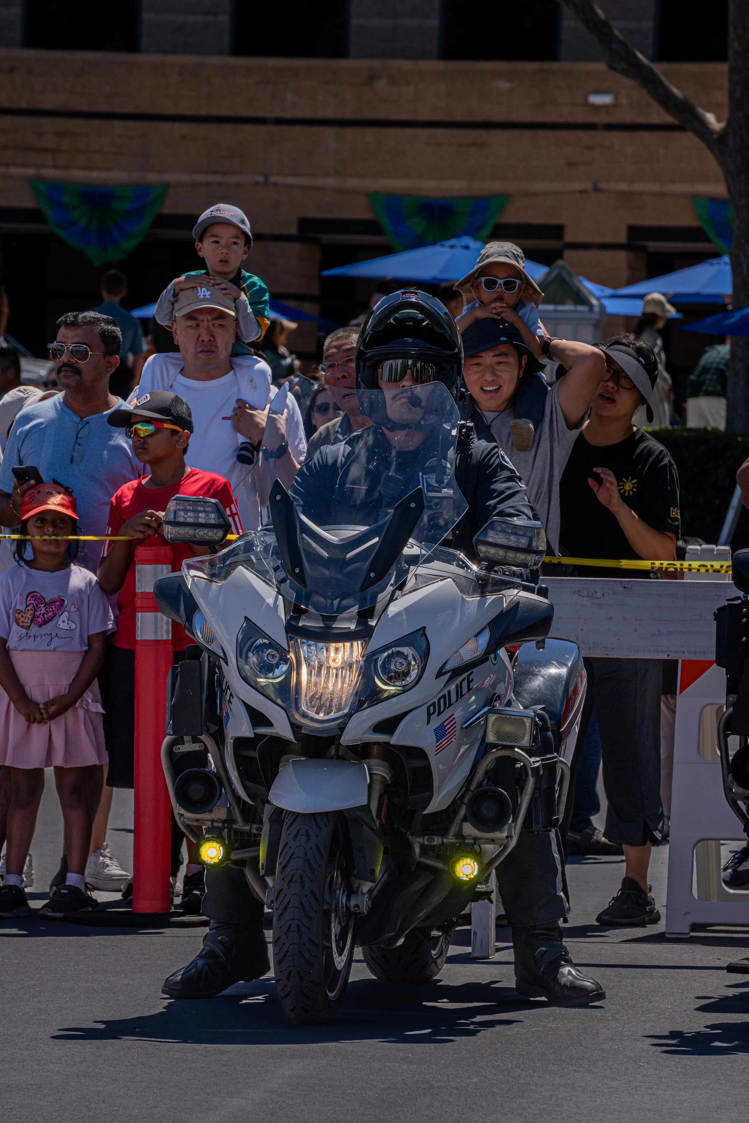 A police officer riding a police motorcycle through a crowd at an outdoor event. The crowd includes children and adults, with some taking photos or videos.