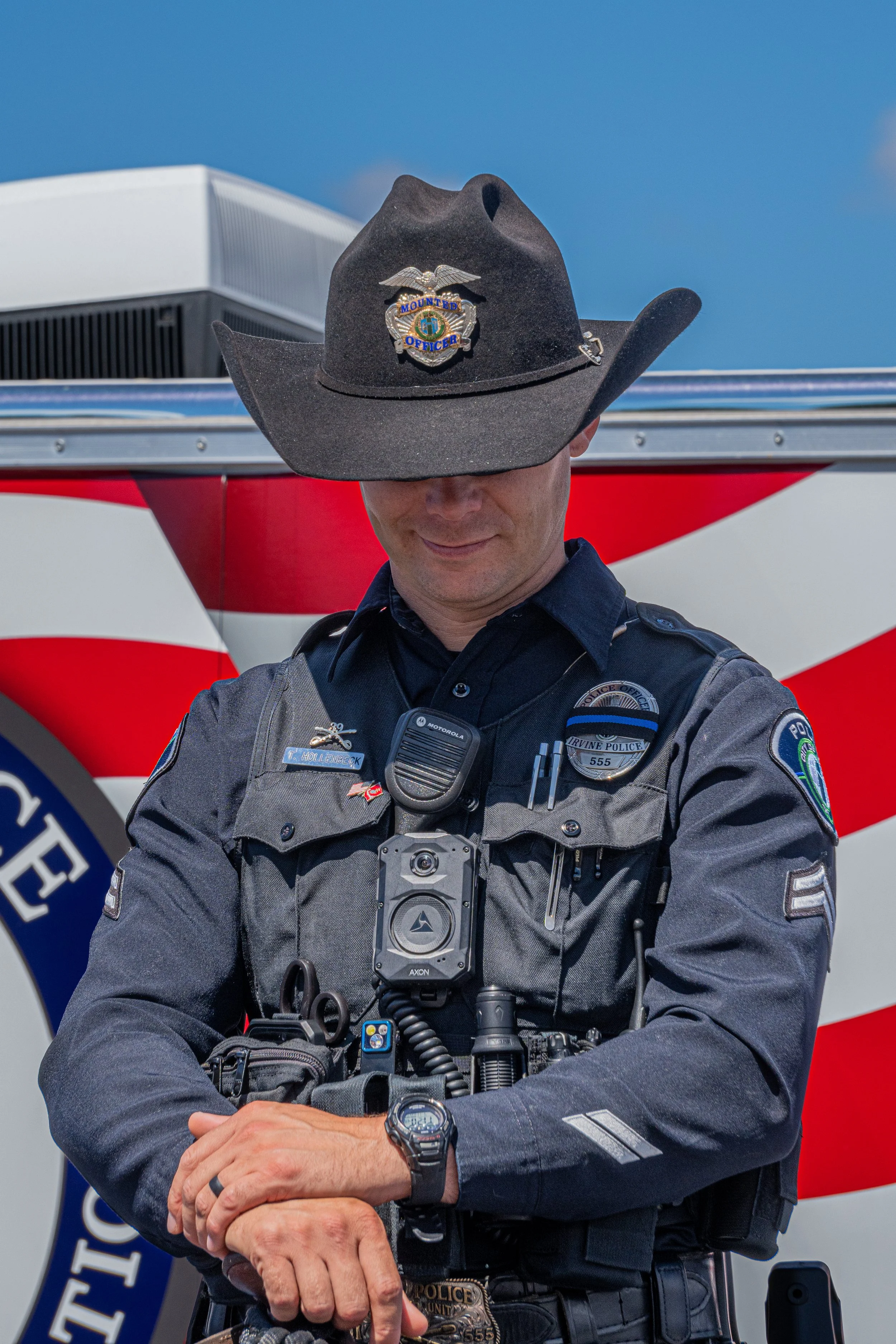A police officer is standing in front of a fire truck, wearing a cowboy hat that has a badge on it. The officer is in uniform with various equipment and a body camera, and is looking down with a slight smile.