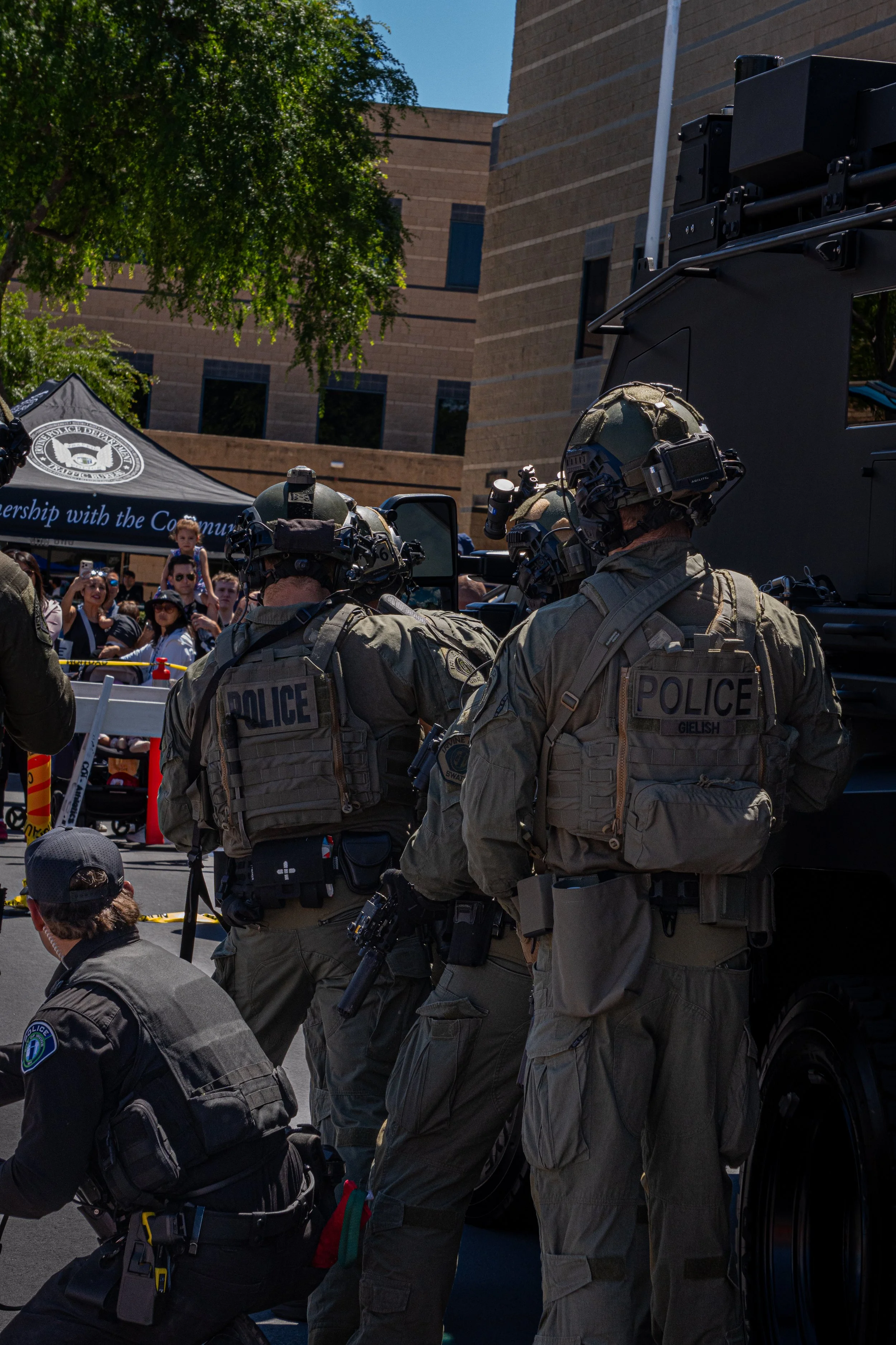 Group of police officers in tactical gear standing near a vehicle at a public event with spectators in the background.