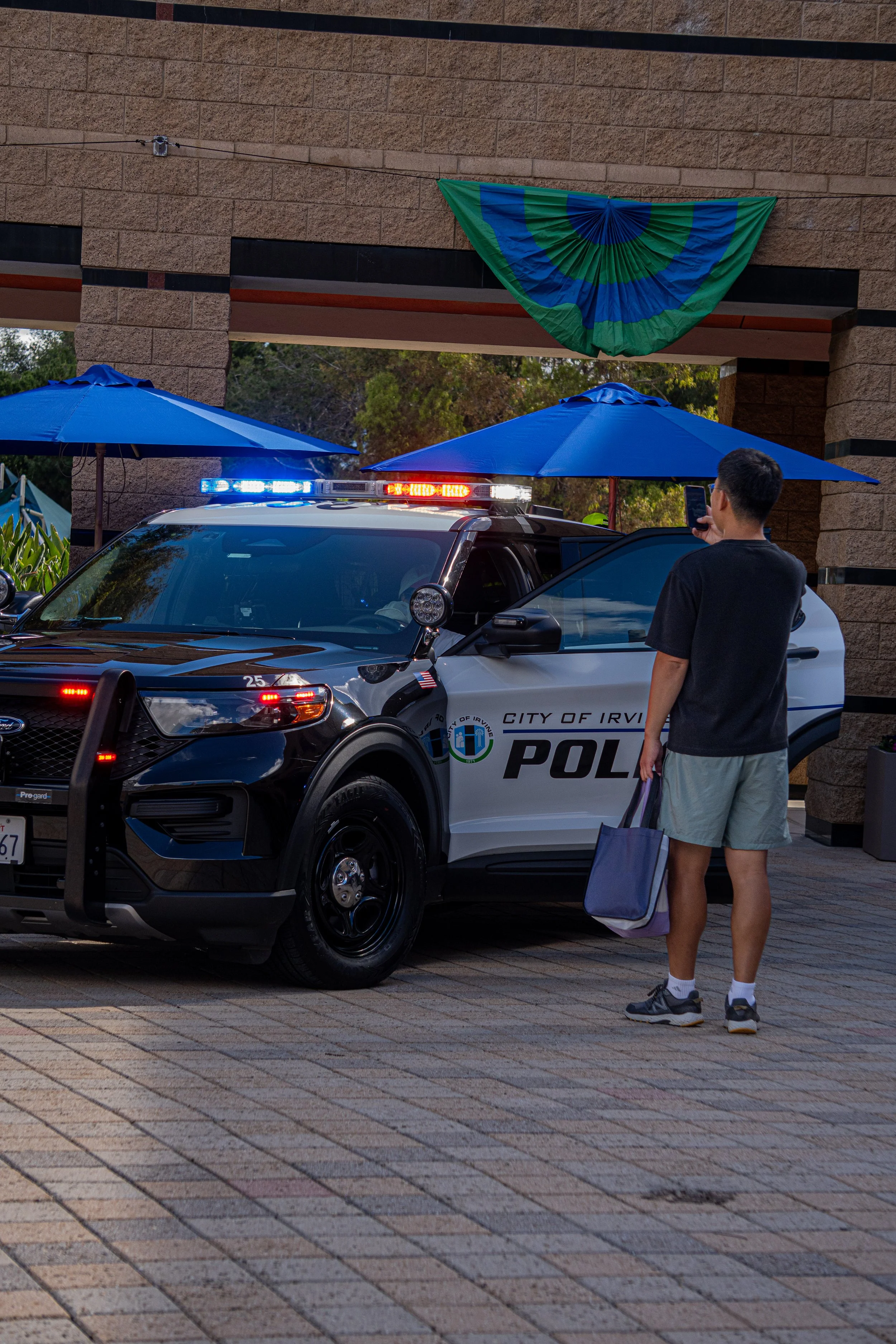 A person standing next to a police car taking a picture with their phone. The police car belongs to the City of Irvi police department and has flashing emergency lights on. The scene is outdoors with blue umbrellas and a decorative green and blue fan