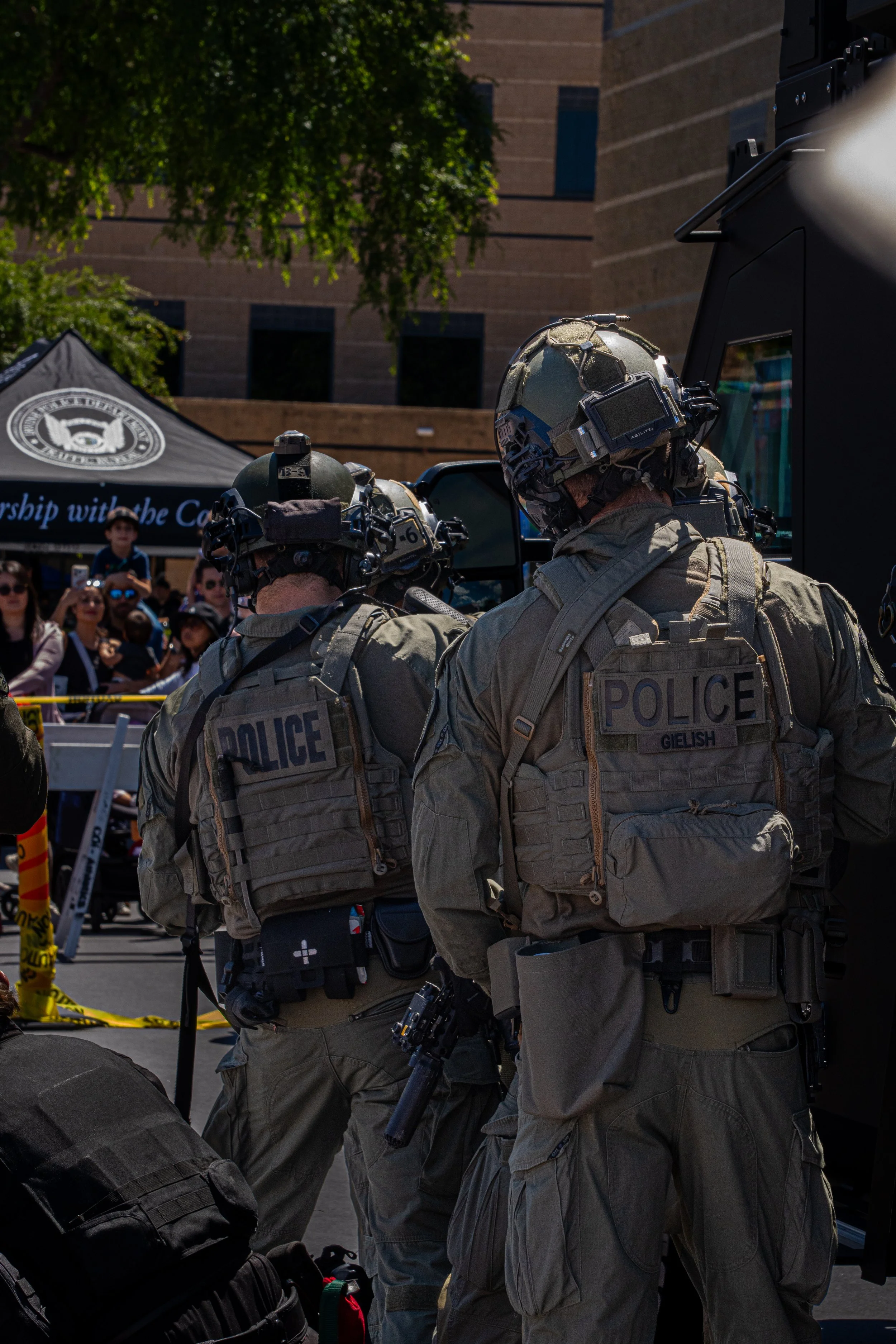 Two police officers in tactical gear and helmets at an outdoor event with a crowd of onlookers and a tent in the background.