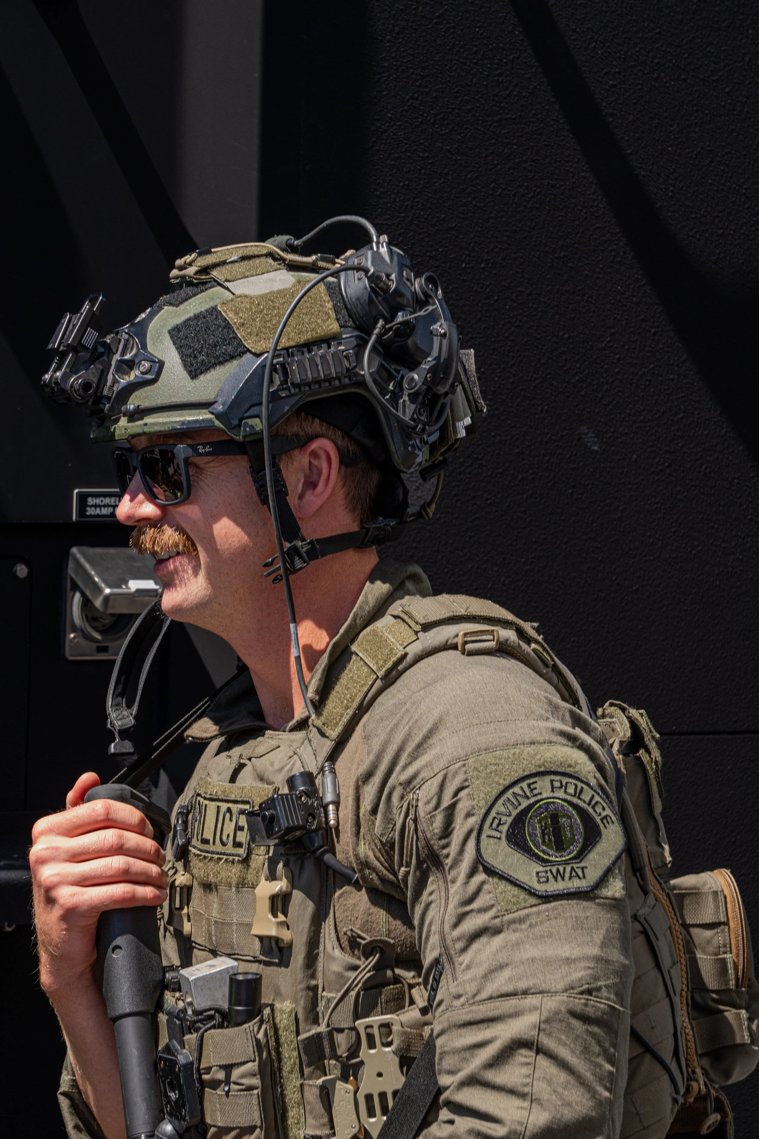 A military police officer in tactical gear wearing a helmet with various attachments and sunglasses, smiling and holding a baton.