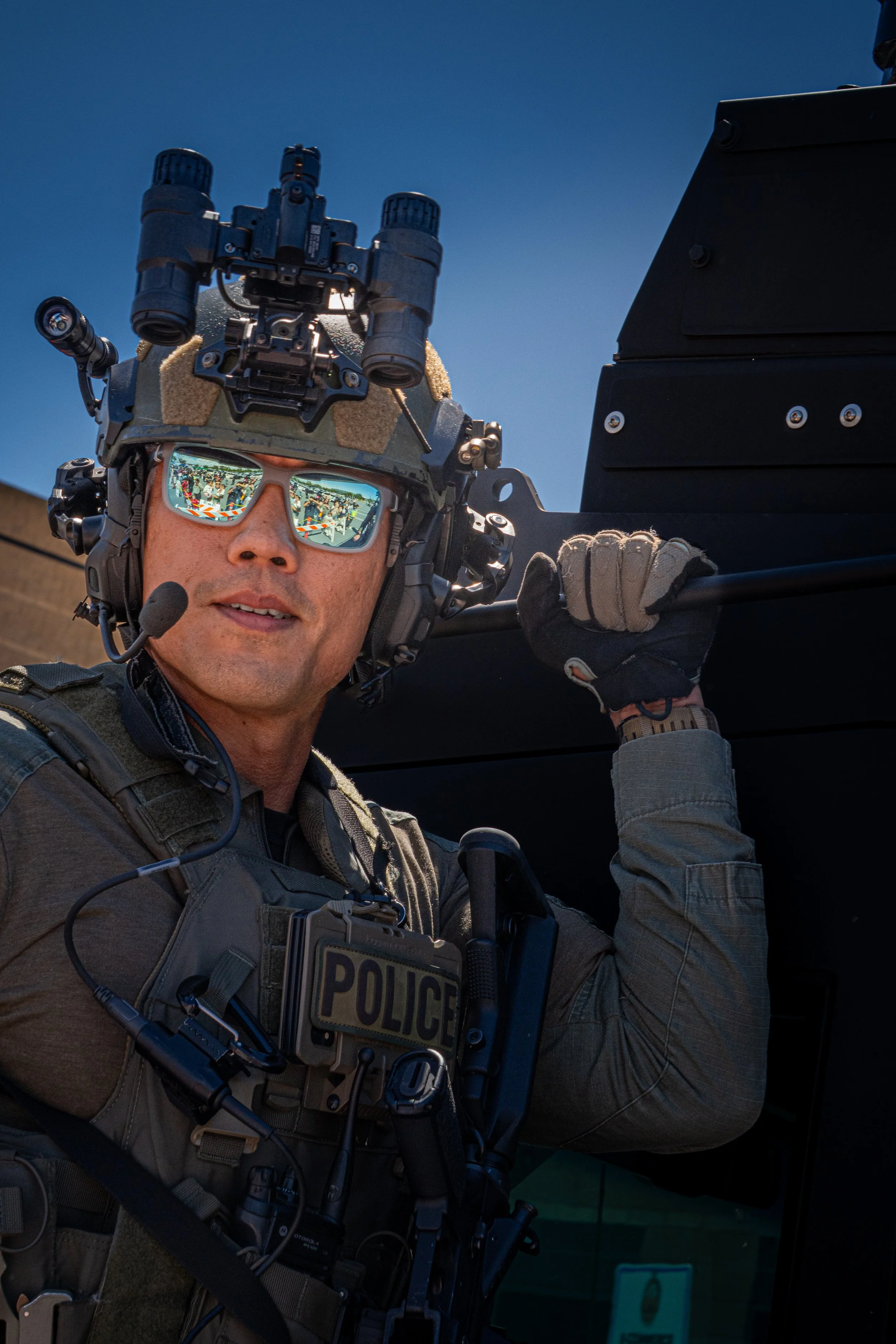 A police officer in tactical gear, wearing sunglasses and a helmet with night vision goggles, posing outdoors against a blue sky.