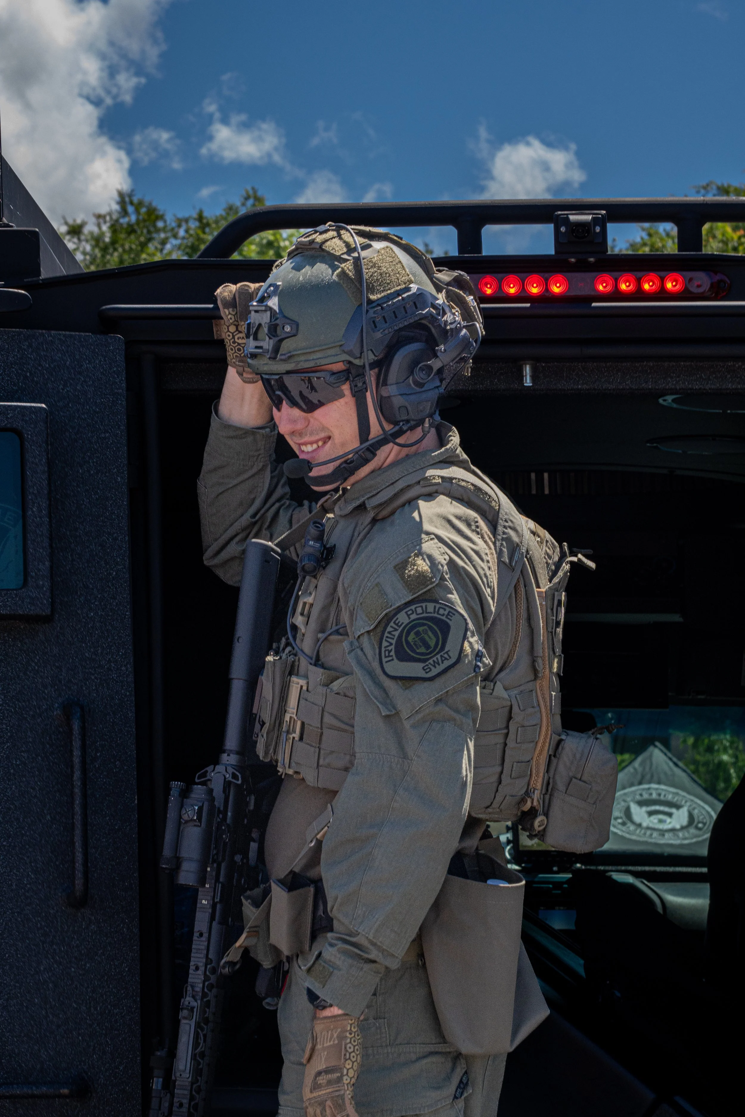 A police officer in tactical gear smiling next to a police vehicle during daytime.