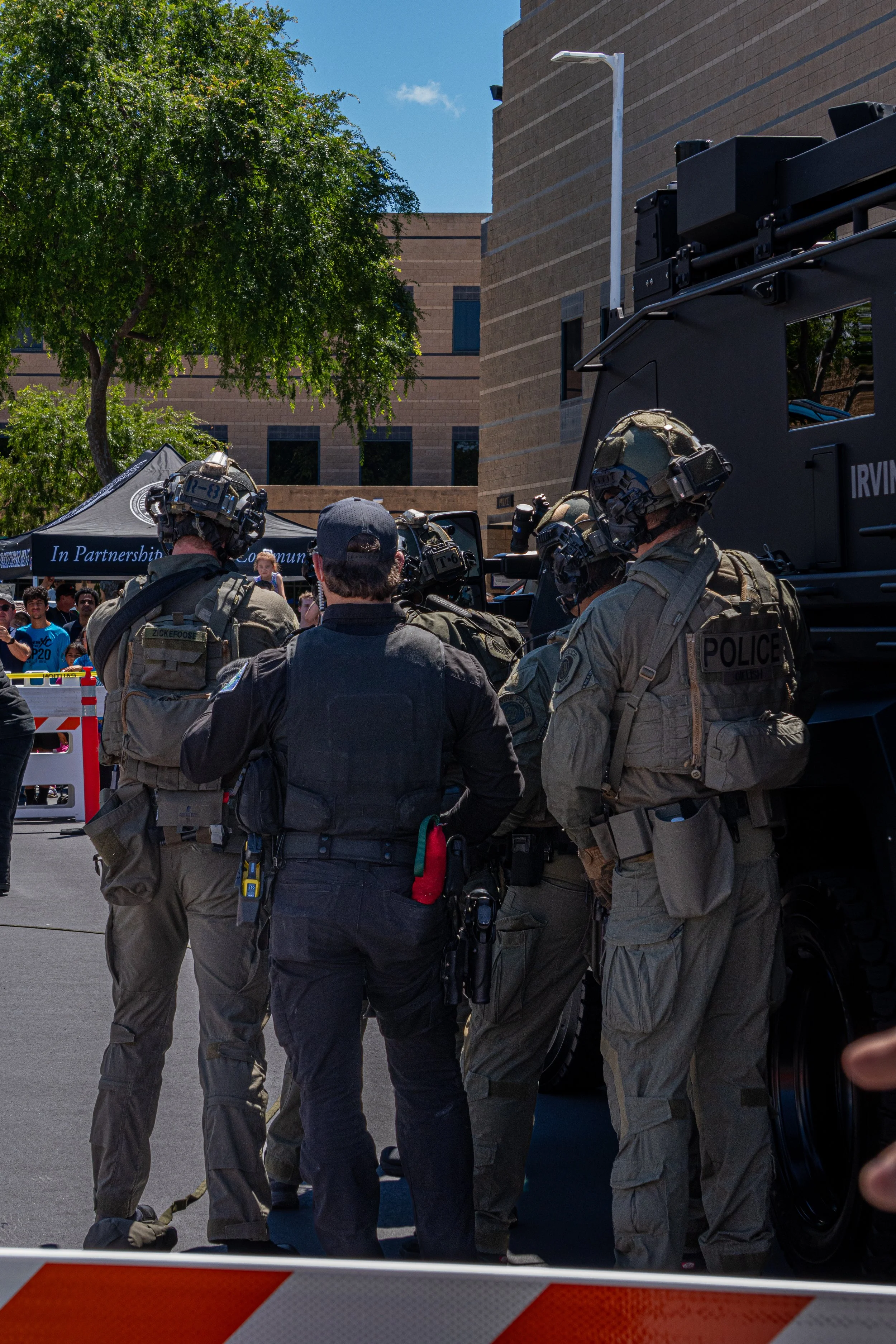 A group of police officers in tactical gear and helmets gather around a police vehicle during a public event.