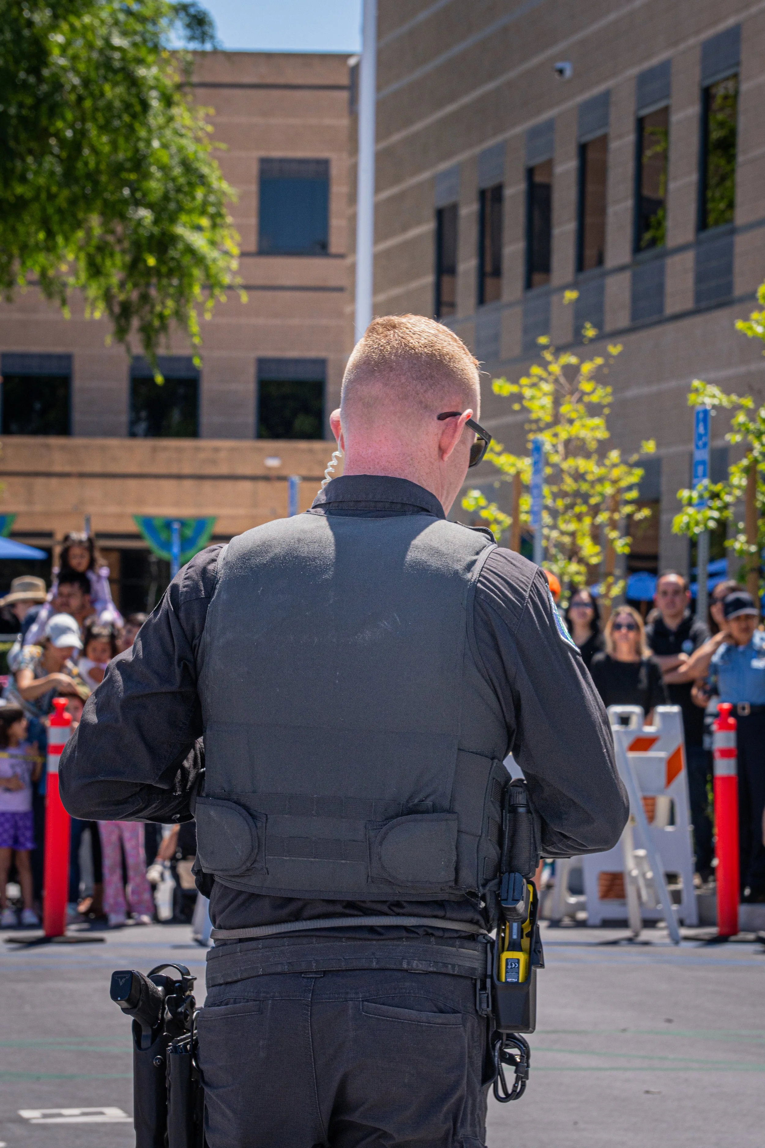 A police officer with short hair, wearing sunglasses and a black uniform, stands with his back to the camera in front of a crowd at an outdoor event on a sunny day.
