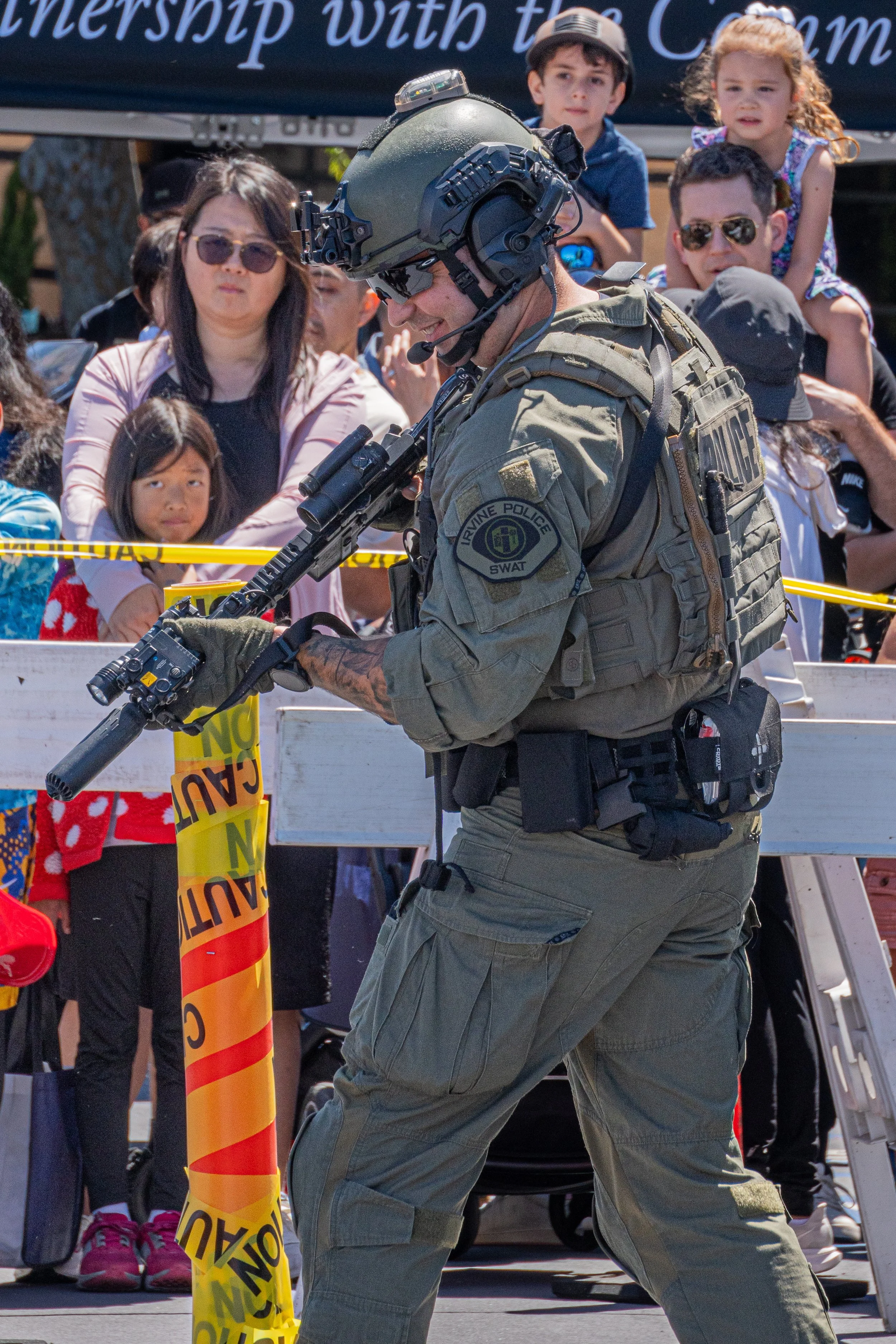 A police officer in tactical gear at a public event, holding a rifle, surrounded by a crowd of children and adults behind police caution tape.