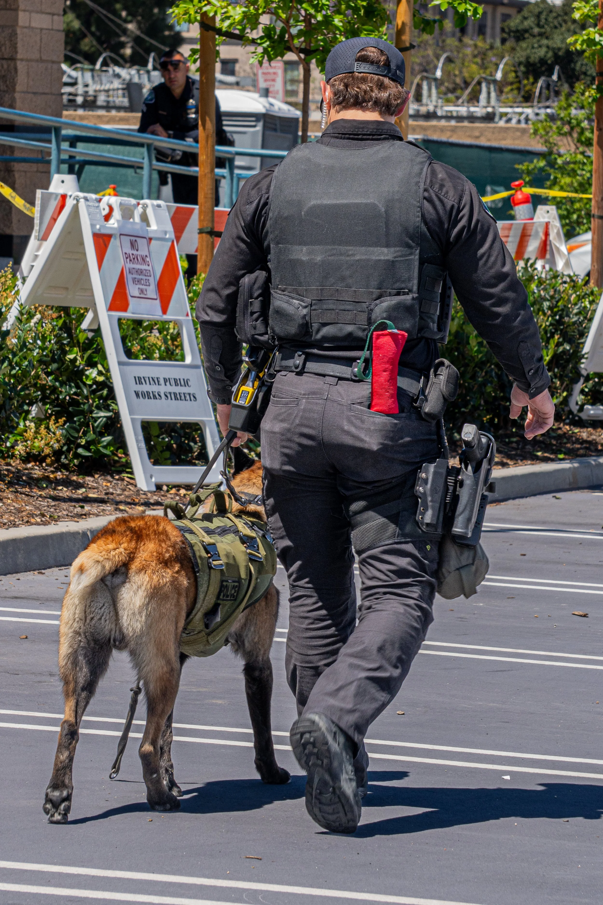 A police officer walking with a police dog in a parking lot near street barriers and a sidewalk, with a person standing in the background.
