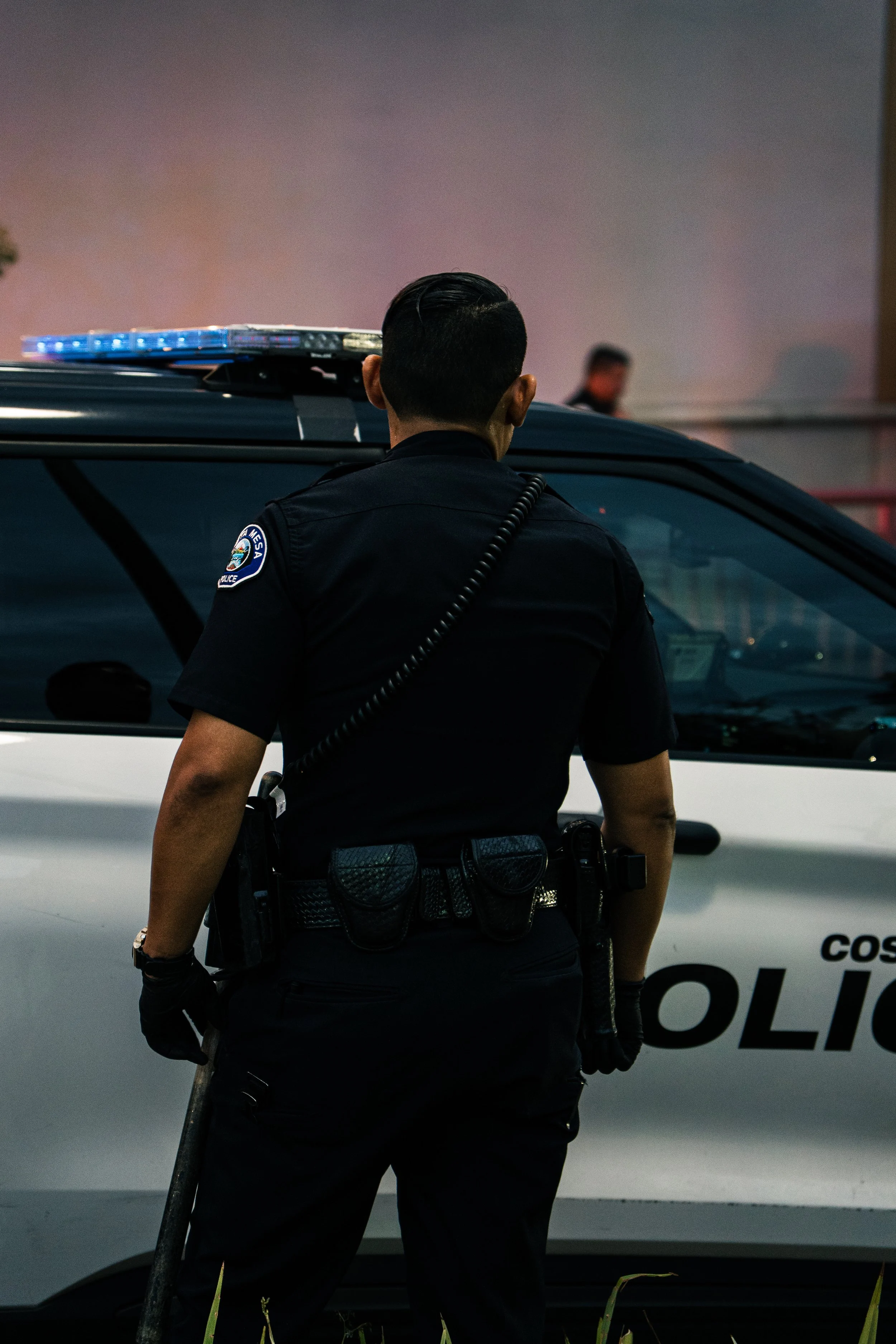 A police officer with short dark hair, wearing a black uniform with a badge, stands in front of a police vehicle at dusk or night, with blurred figures in the background.