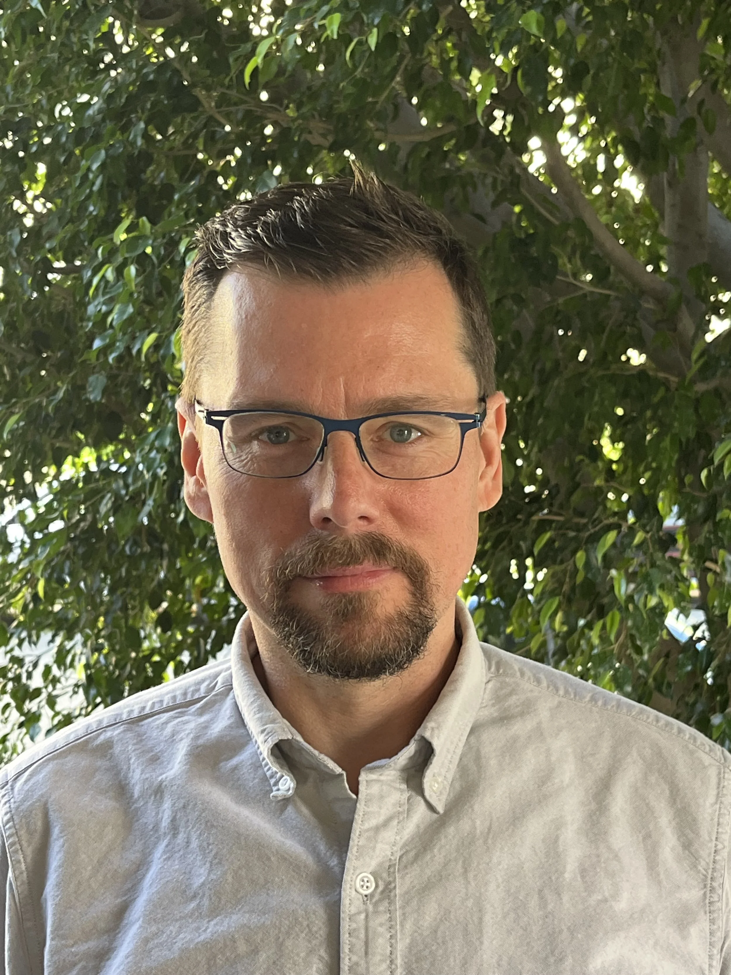 A man with short dark hair, glasses, and a goatee, wearing a light-colored collared shirt, standing outdoors in front of a leafy tree with sunlight filtering through the branches.