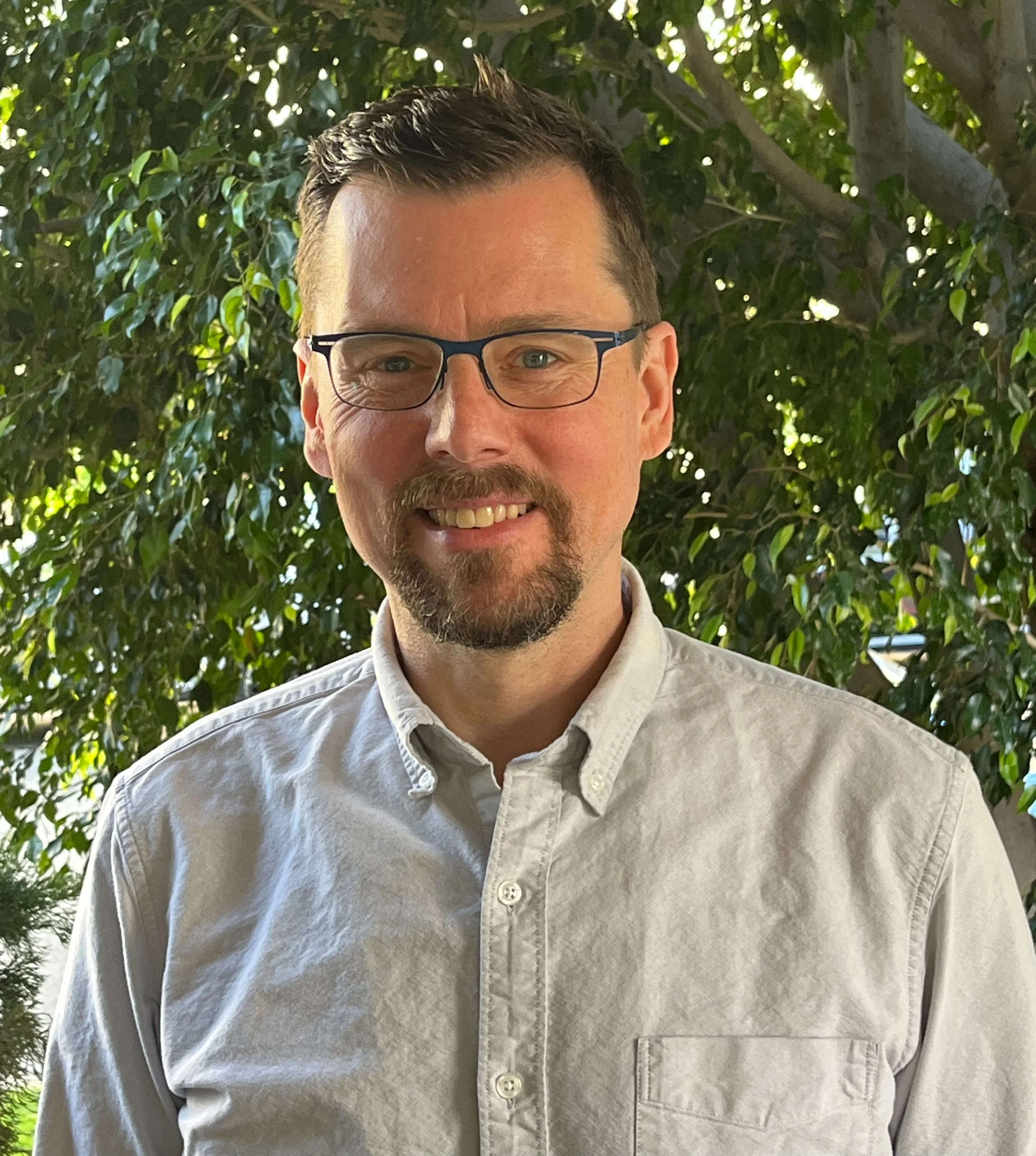 A smiling man with glasses and a beard, wearing a light-colored button-up shirt, standing outside in front of a large leafy green tree.
