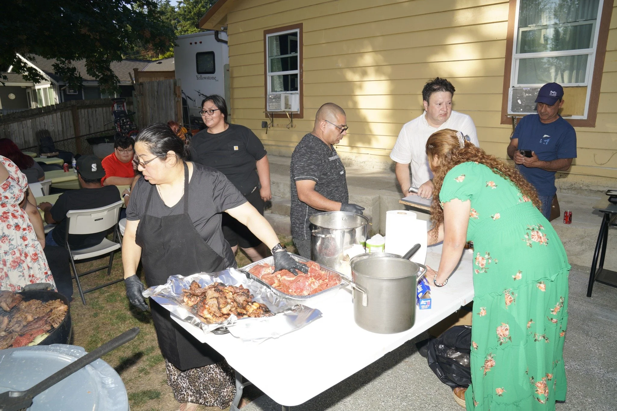 People gather around a table with food at an outdoor barbecue party in a backyard with a yellow house in the background.