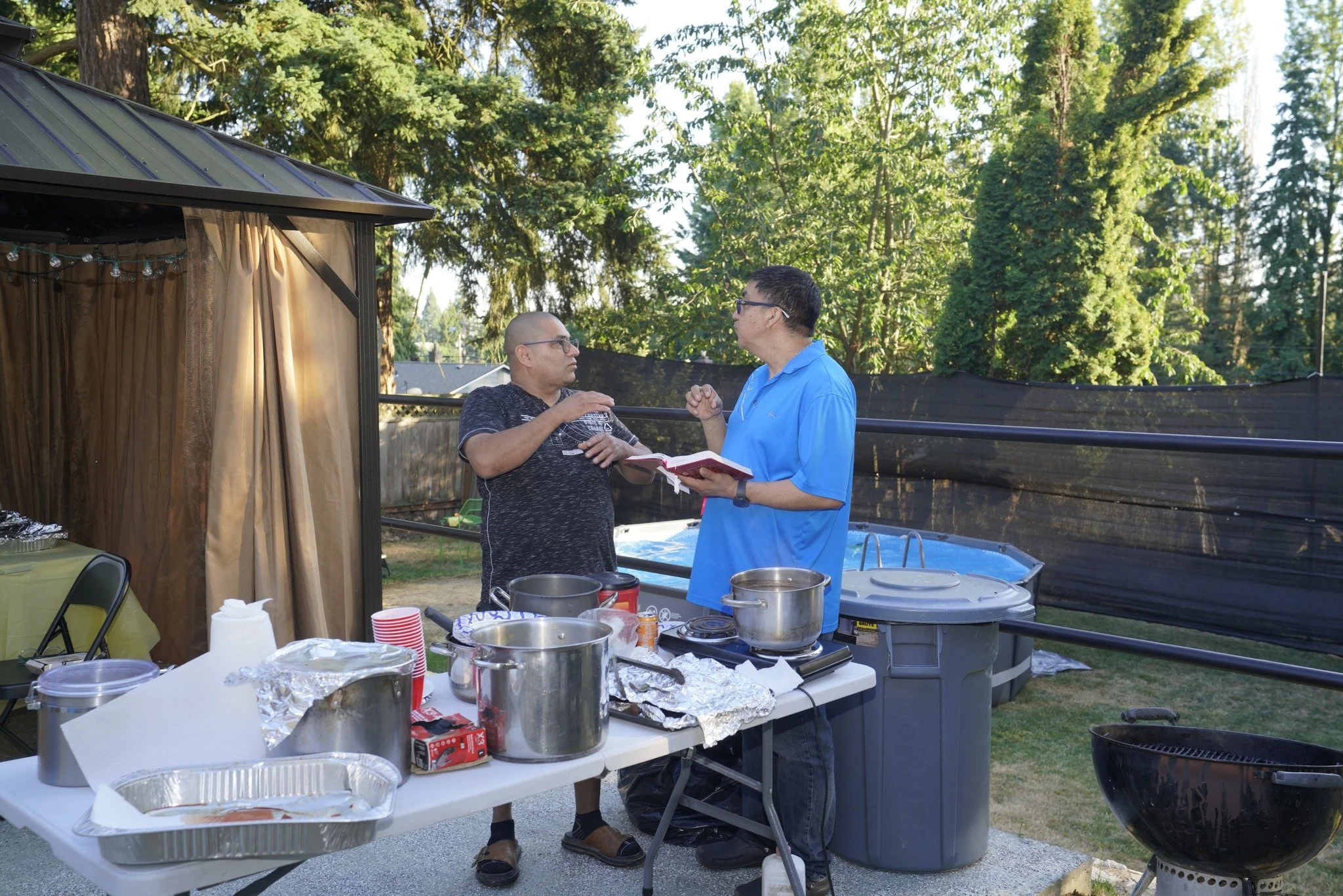 Two men having a conversation at a backyard barbecue with a table of food and cooking supplies nearby, surrounded by trees and a black privacy fence.