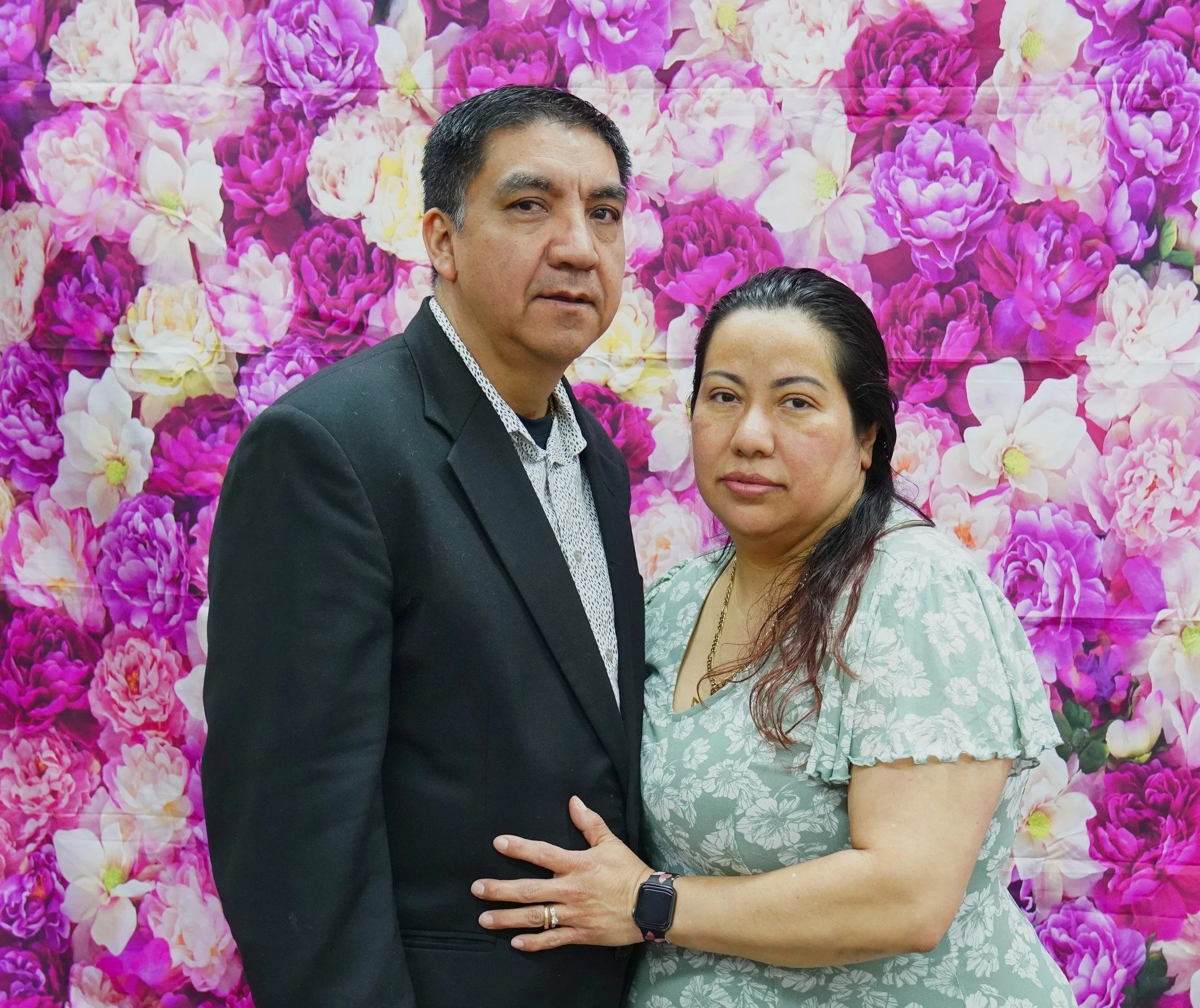 A middle-aged man in a dark suit and dress shirt standing next to a woman in a light floral dress, against a backdrop of pink and purple peony flowers.