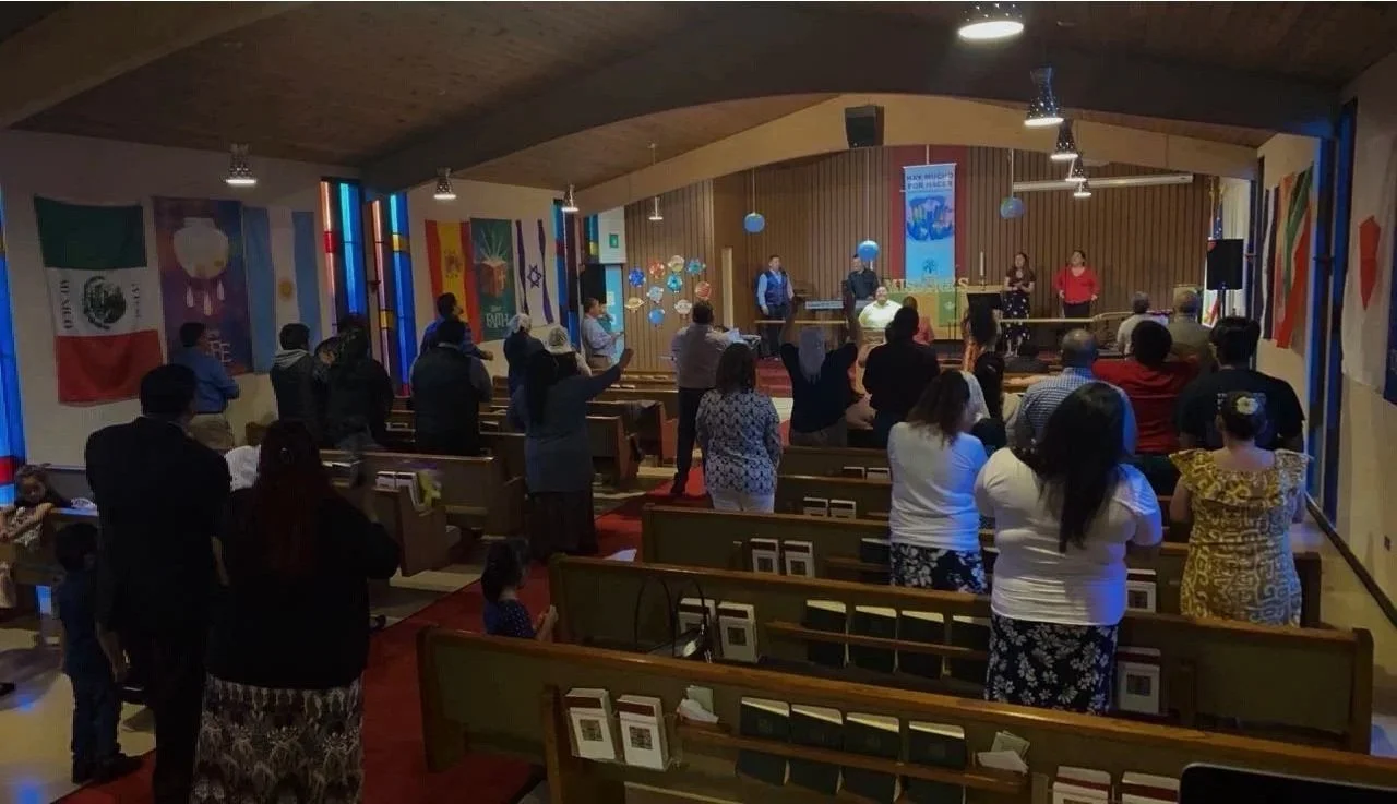 People gathered in a church or community hall, standing and raising hands towards a stage where a group of women and a man are leading a service or performance, with colorful banners and flags hanging on the walls.