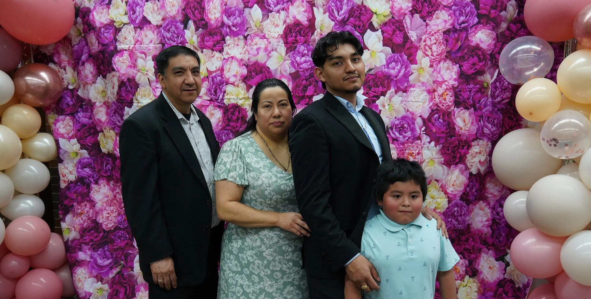 Family members standing in front of a pink floral backdrop with balloons, posing for a photo at a celebration or party.