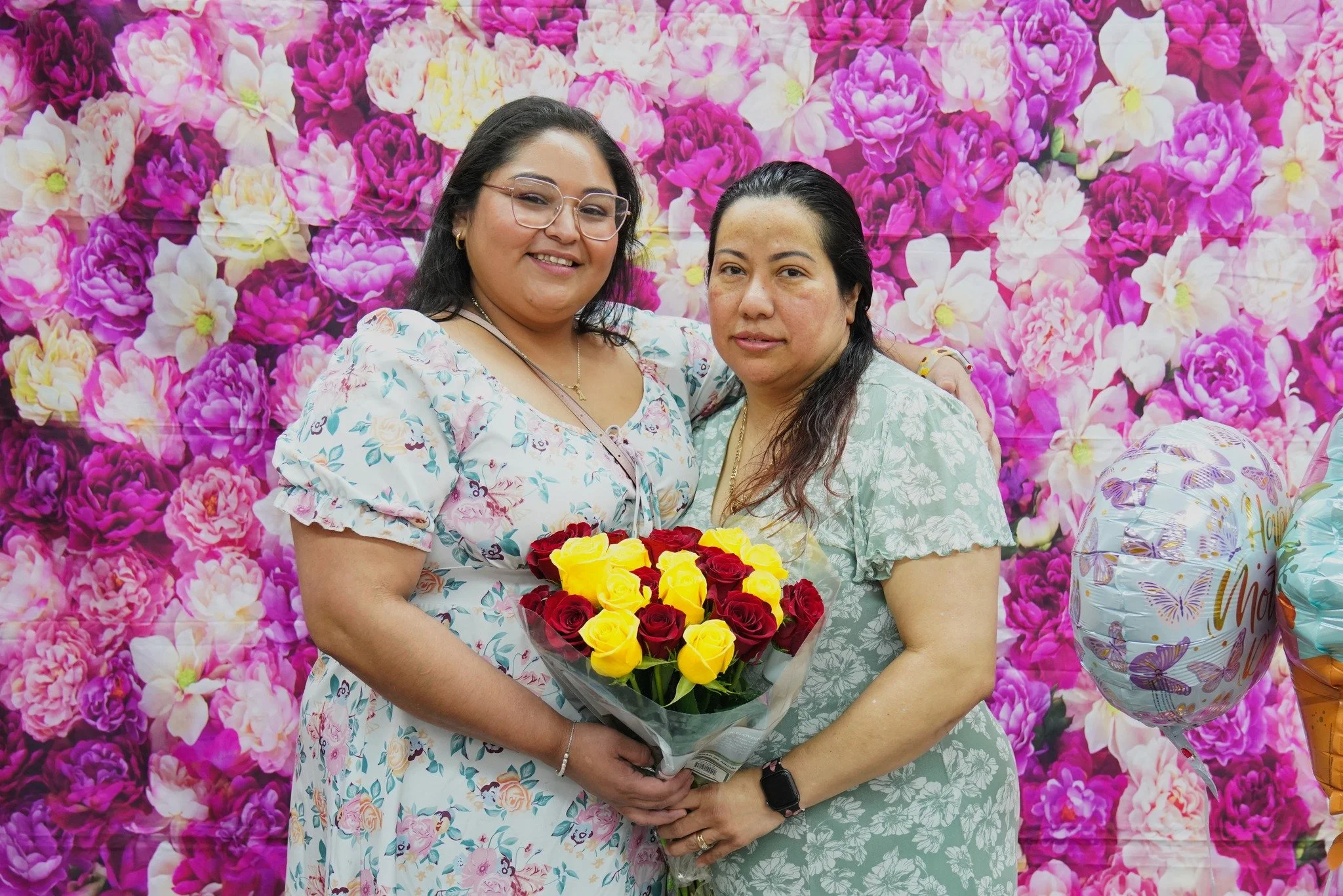Two women standing close together in front of a pink floral backdrop, holding a bouquet of red and yellow roses, celebrating Mother's Day.