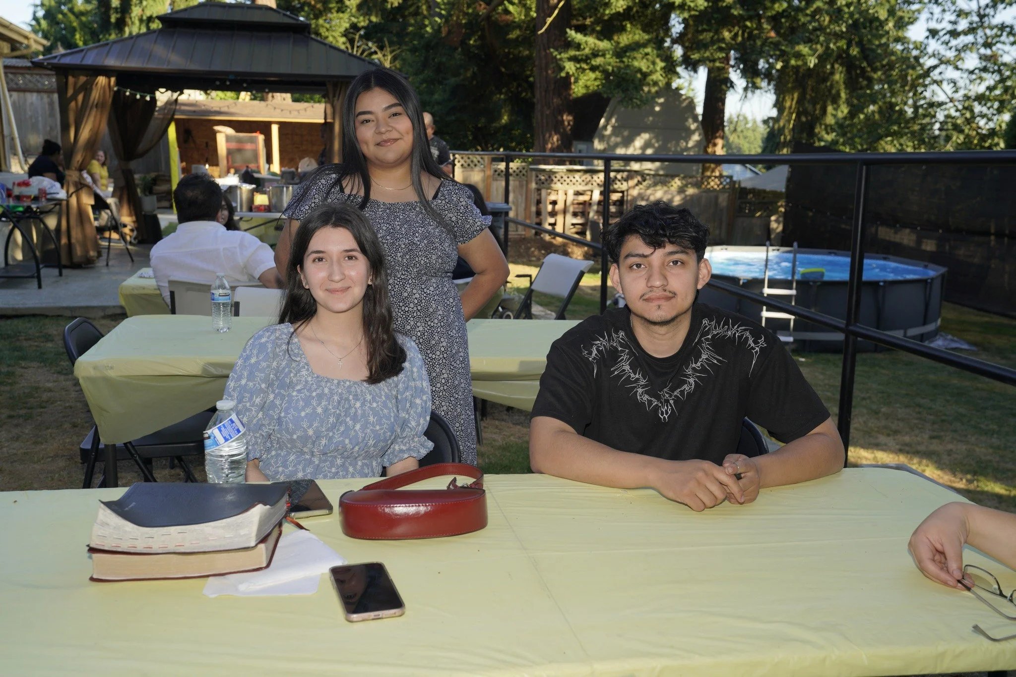 Three teenagers sitting and standing at a yellow-covered table outdoors during daytime, with other people and a hot tub in the background.