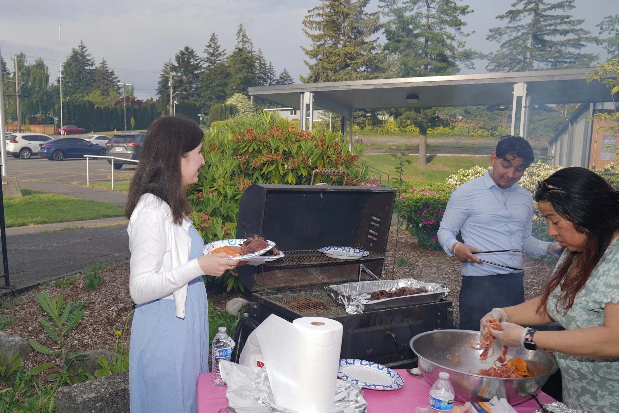 Three people preparing food at an outdoor barbecue. One woman standing by the grill holds a plate of cooked meat, while another woman uses tongs to handle food in a large bowl. A young man stands near her, possibly serving or preparing to cook additi
