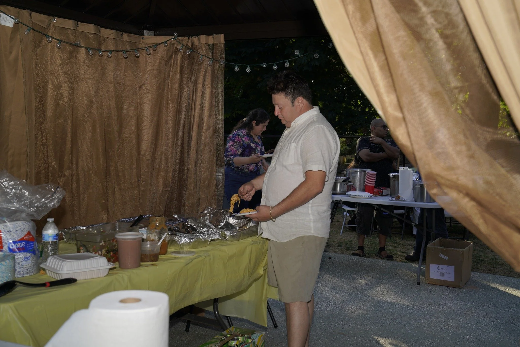 A man in a white shirt and beige shorts is serving himself food at an outdoor buffet with a yellow tablecloth, under a canopy with brown curtains and string lights, during a gathering. In the background, two women are near a table with supplies.