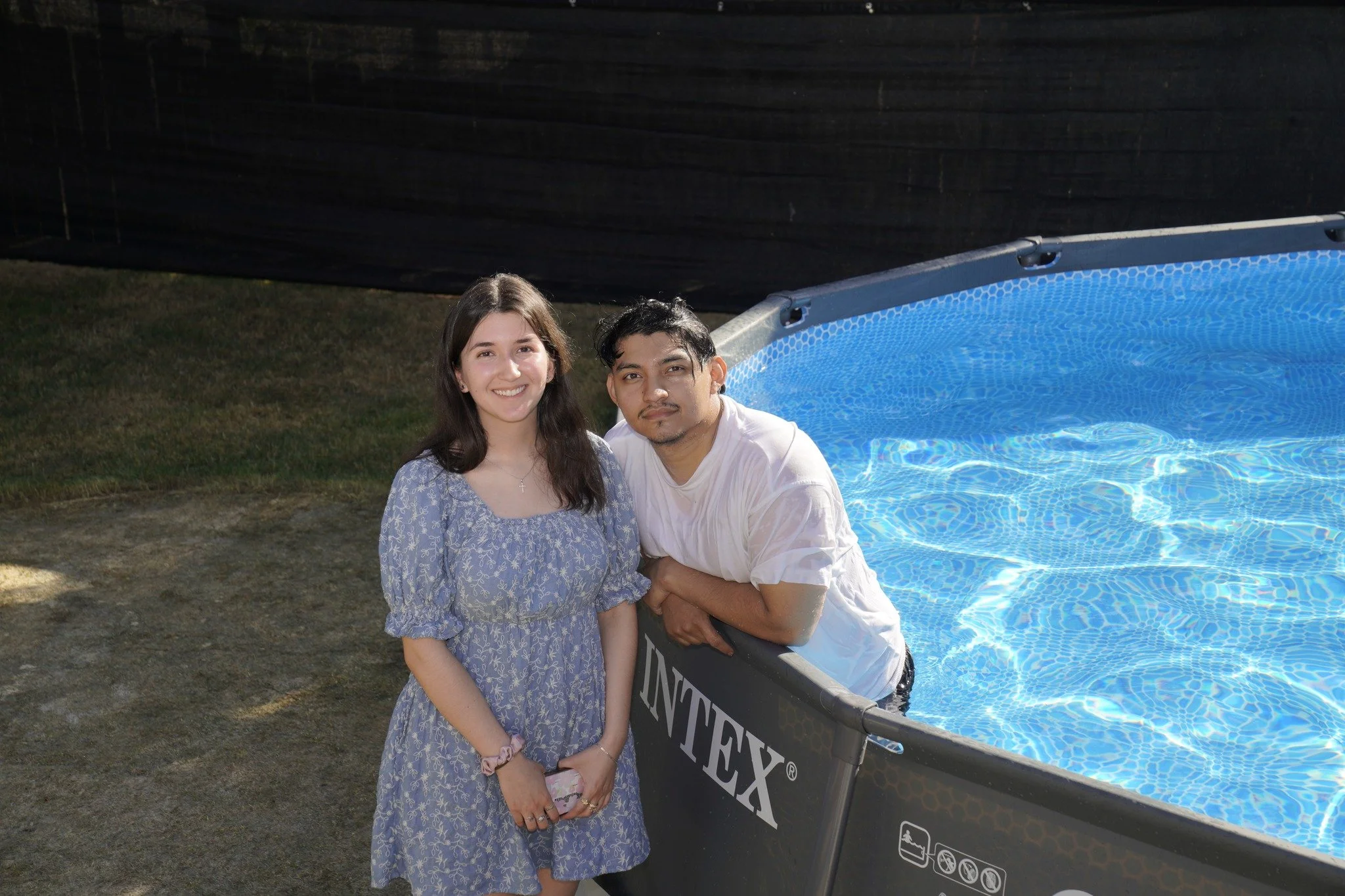 A young woman and a young man are posing next to a blue above-ground swimming pool outdoors in the daytime. The woman is smiling, wearing a blue dress, and holding a pink phone. The man is leaning on the pool, wearing a white t-shirt, and has a neutr