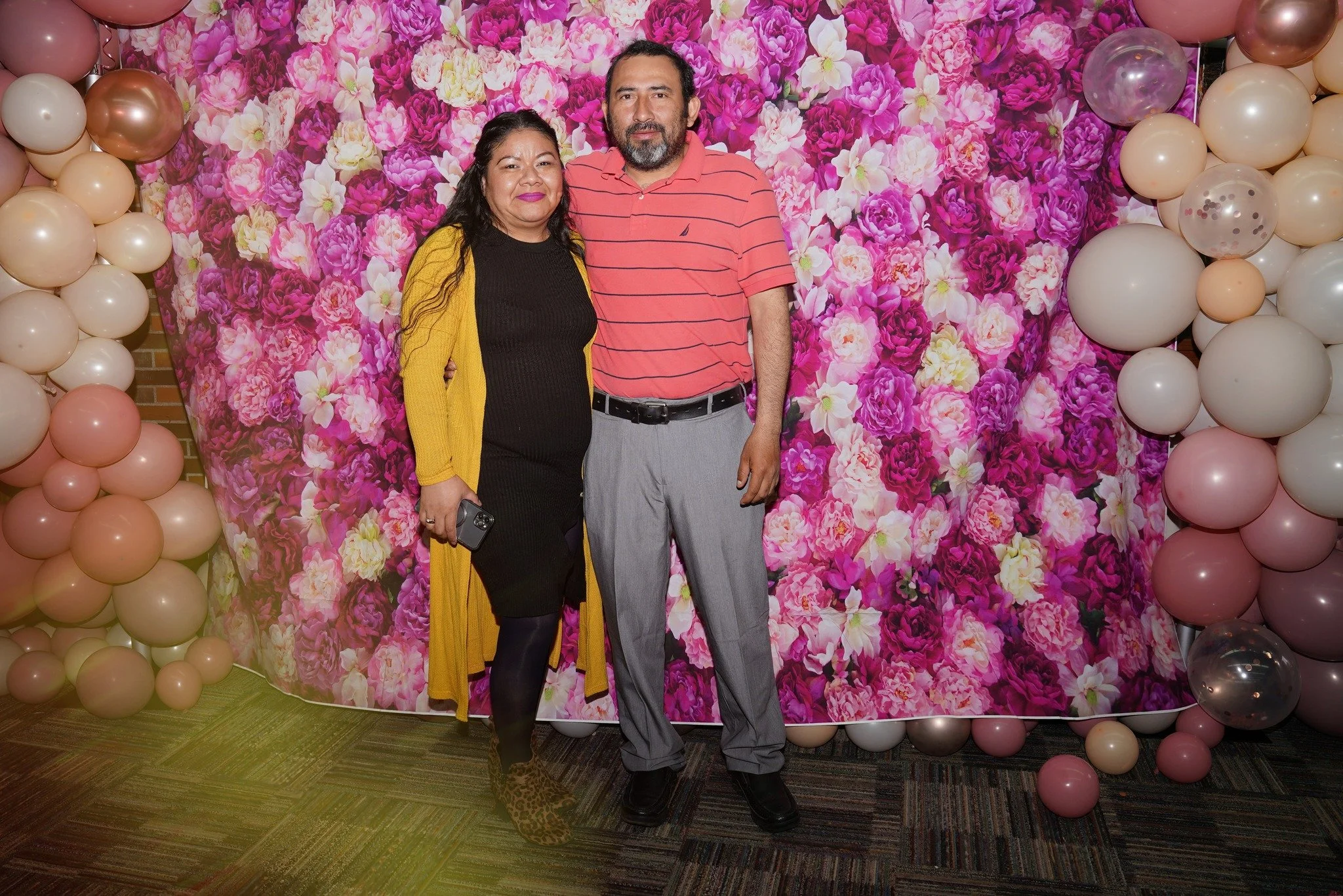 A woman and a man standing together in front of a floral backdrop decorated with pink and purple flowers and balloons.