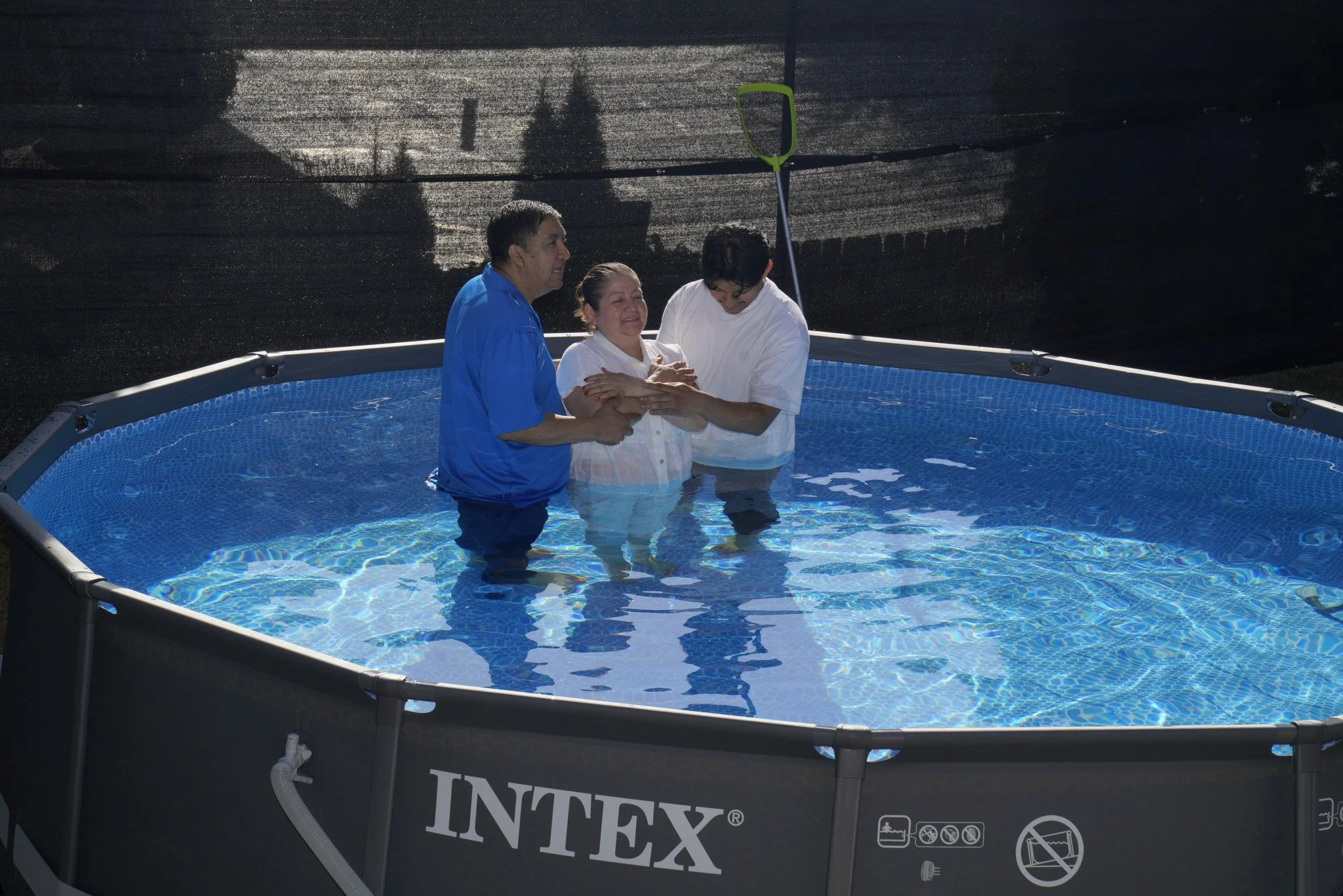 Three people baptizing a woman in a small above-ground pool, with a black privacy screen in the background.