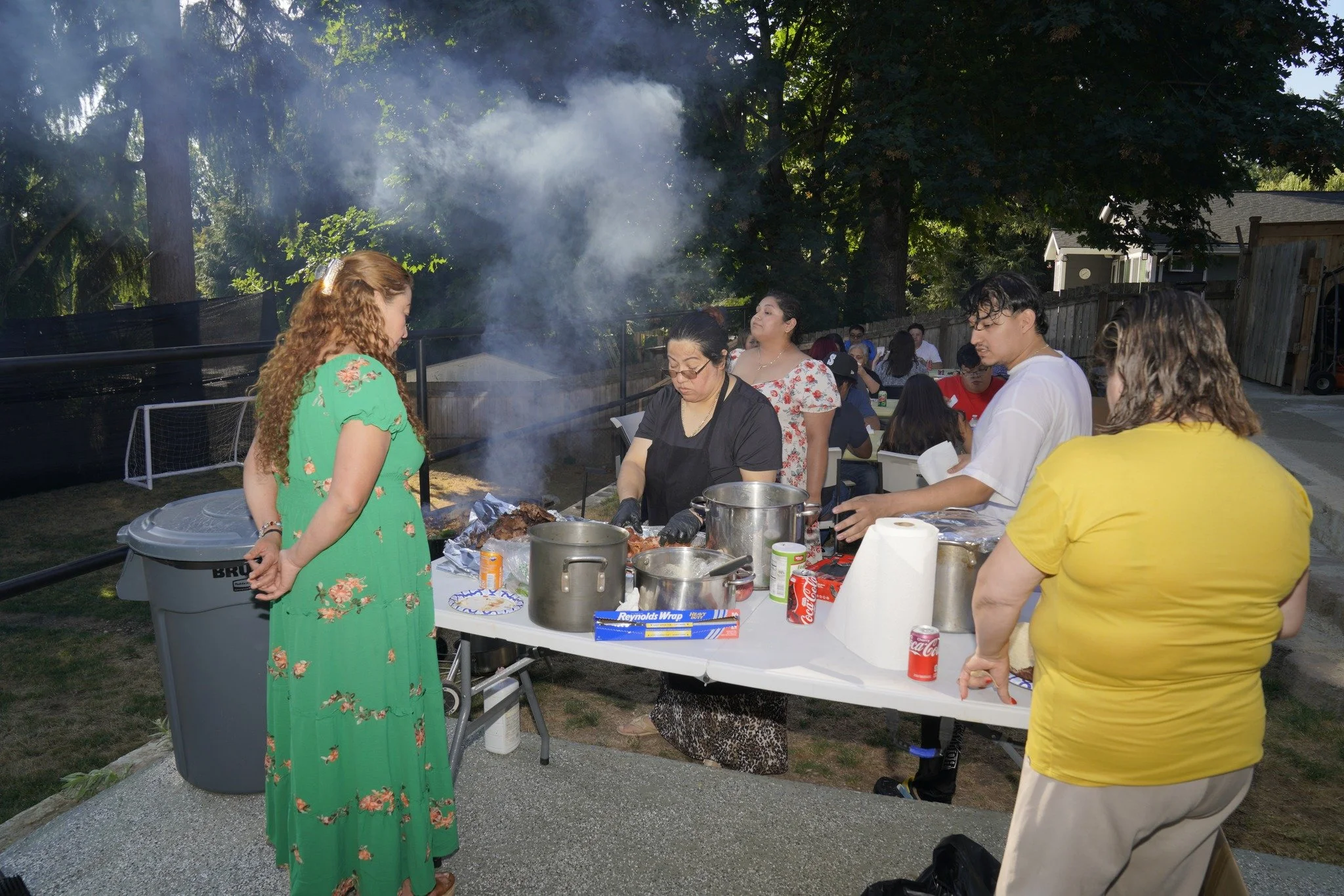 People cooking and serving food outdoors at a gathering, with a grill emitting smoke, on a grassy area with trees in the background.