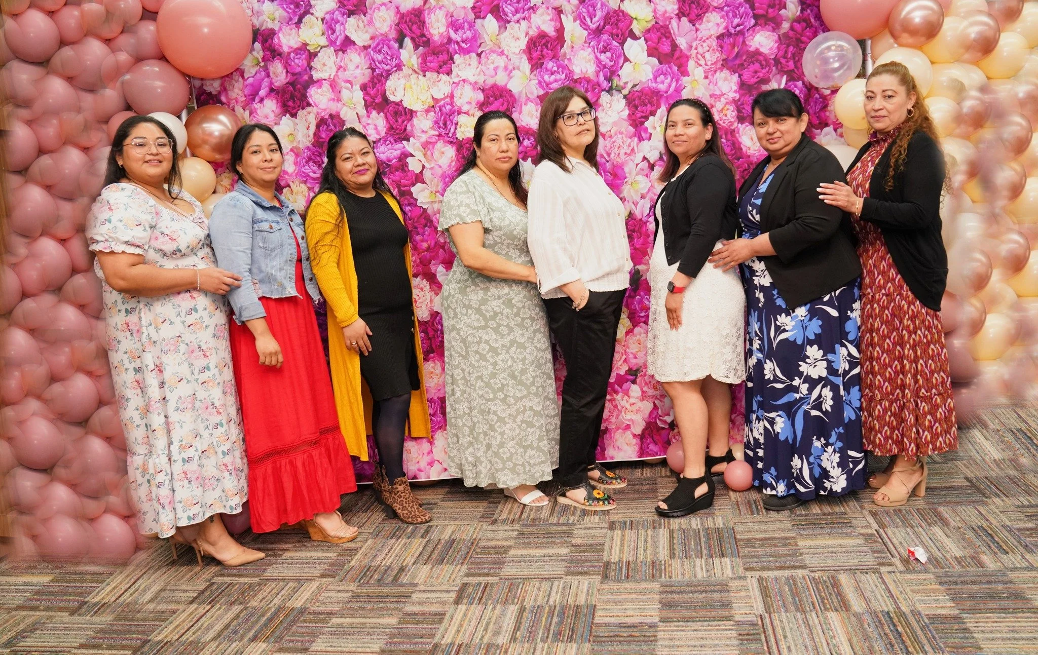 Group of nine women standing in front of a pink floral backdrop with balloons, dressed in colorful dresses, skirts, and jackets, smiling at a celebration or event.