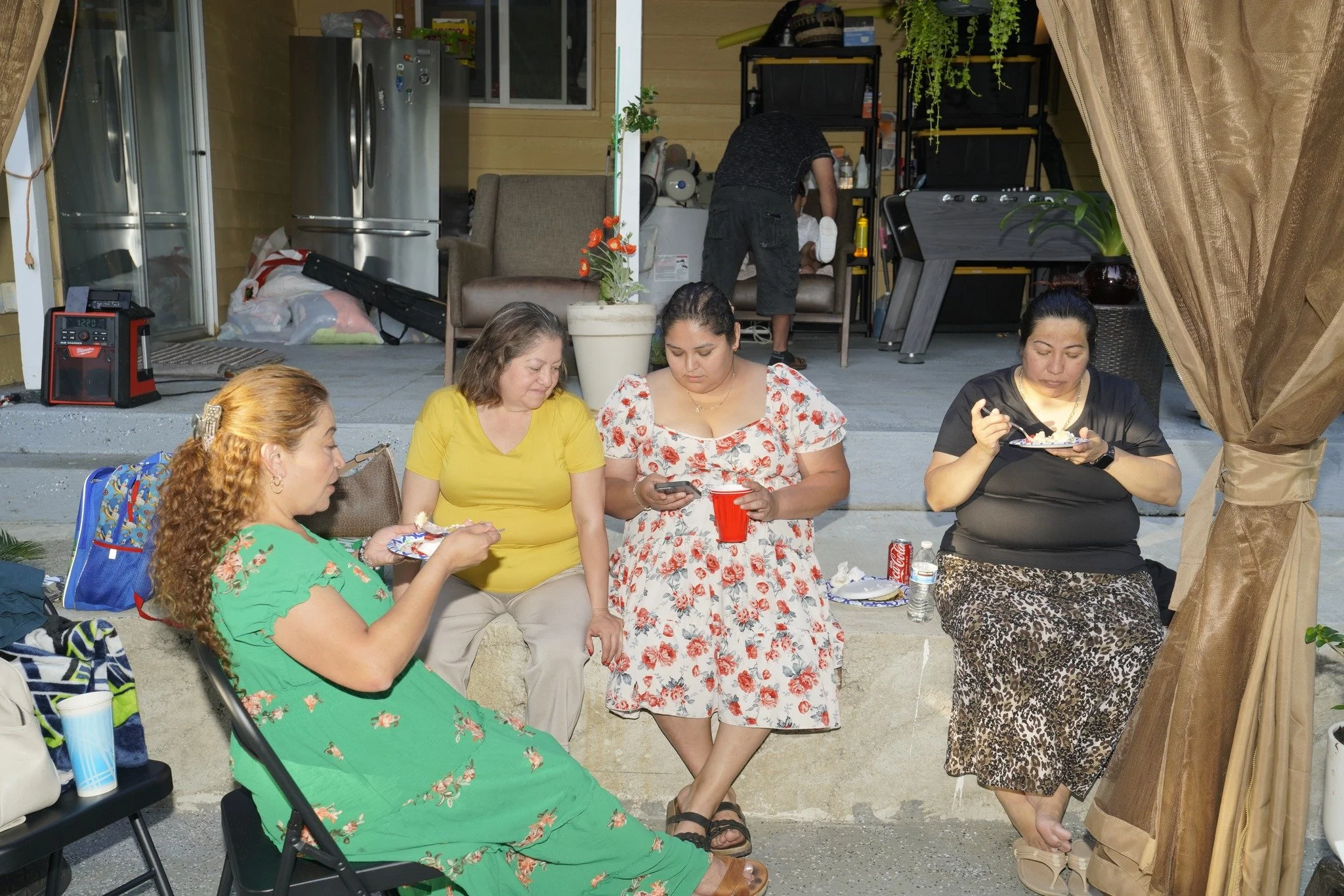 Four women sitting outdoors, eating and using their phones, with a man in the background preparing food.