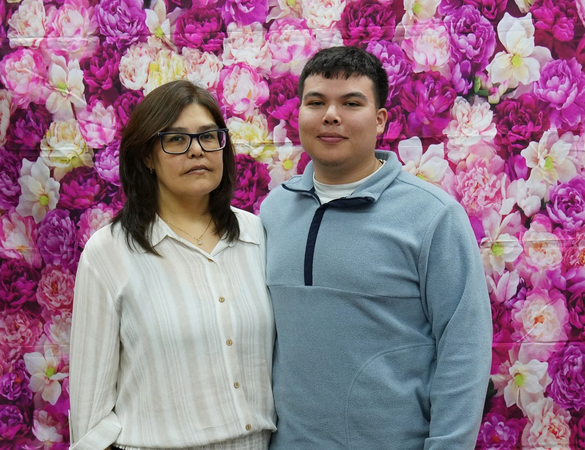 A woman with glasses and a man stand in front of a pink and purple floral background.
