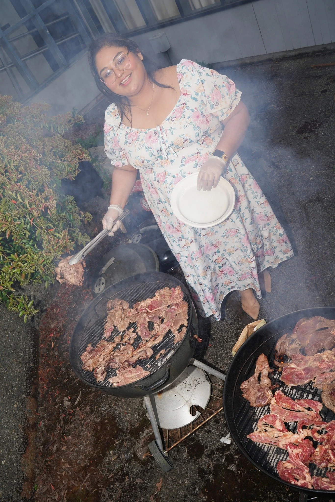 A woman wearing a floral dress and glasses is grilling meat outdoors. She holds tongs with a piece of meat and a plate. Two barbecue grills with cooking meat are in front of her, and she is smiling while cooking.