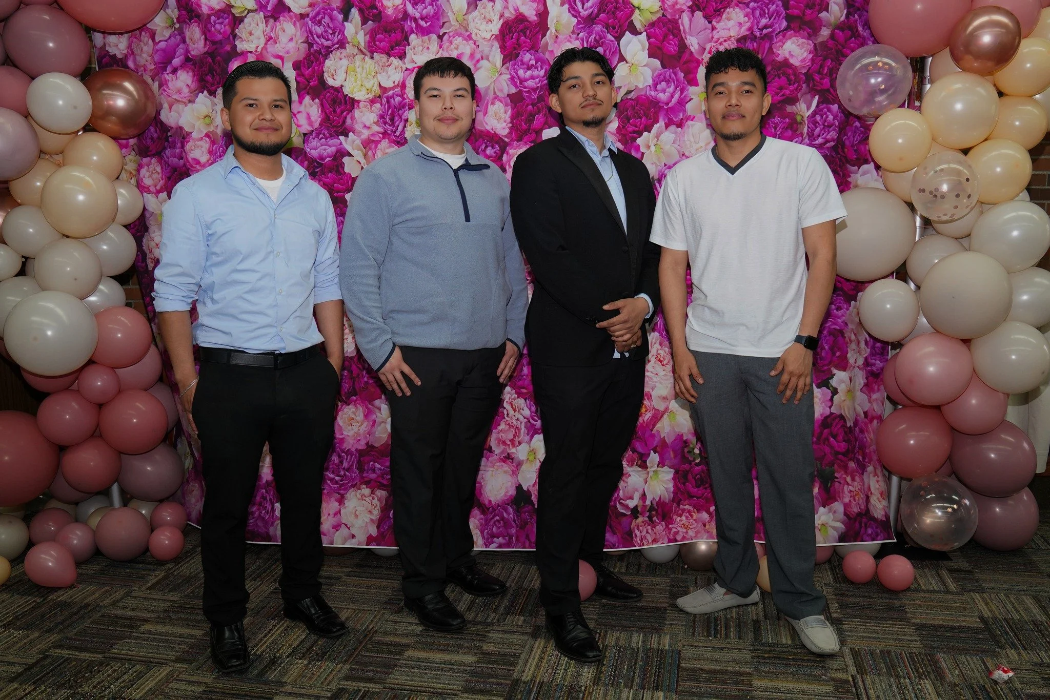 Four young men standing in a row in front of a pink floral backdrop with balloon decorations on either side