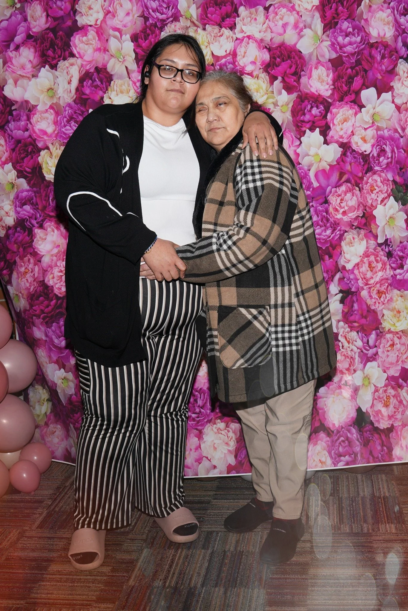 Two women standing close together, embracing each other in front of a pink floral background. One woman is wearing a black cardigan, white shirt, and striped pants; the other is wearing a patterned coat and beige pants.