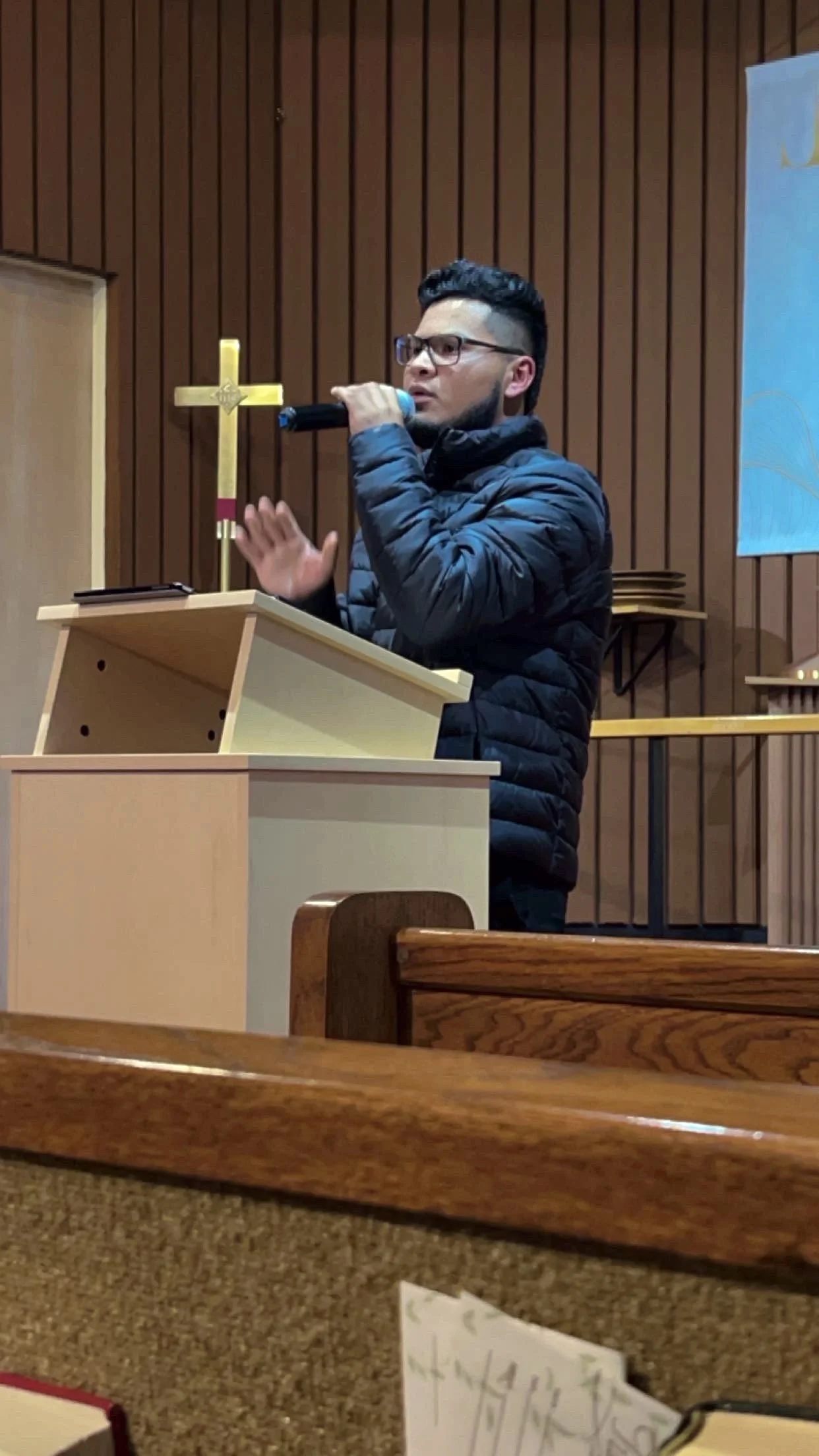 A man wearing glasses and a black puffer jacket standing at a wooden pulpit inside a church, speaking into a microphone. Behind him is a gold cross mounted on a wooden wall.