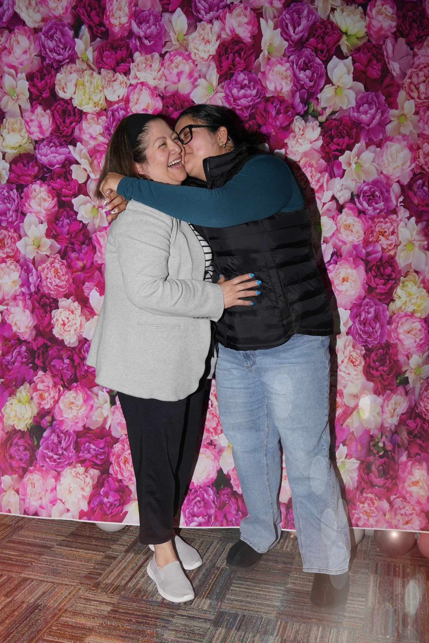 Two women embracing and smiling in front of a vibrant pink floral backdrop, one wearing a gray blazer and the other in a black vest and glasses.